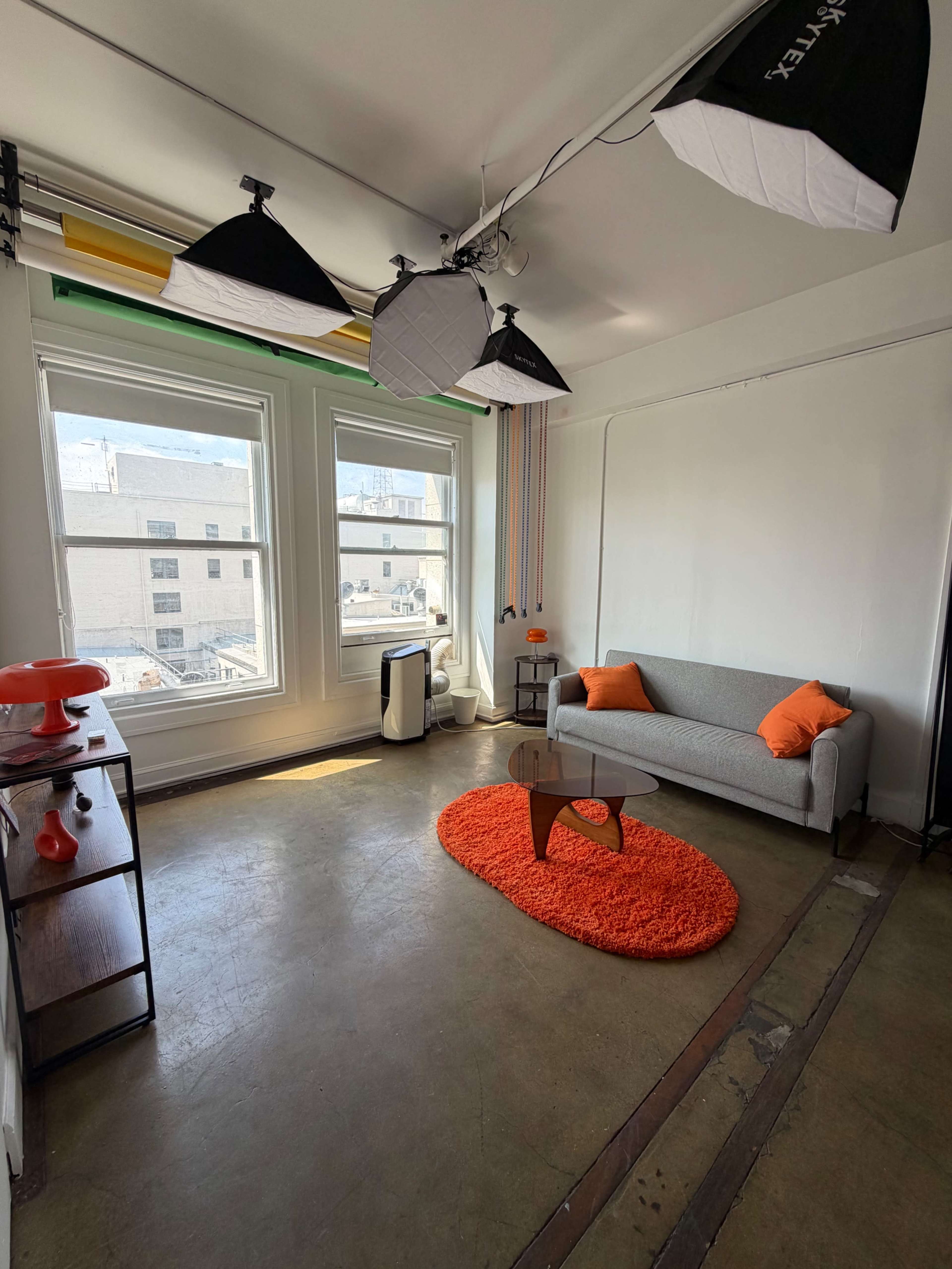 The image shows a bright room with large windows, a gray sofa with orange cushions, a round orange rug, and studio lighting equipment mounted on the ceiling.