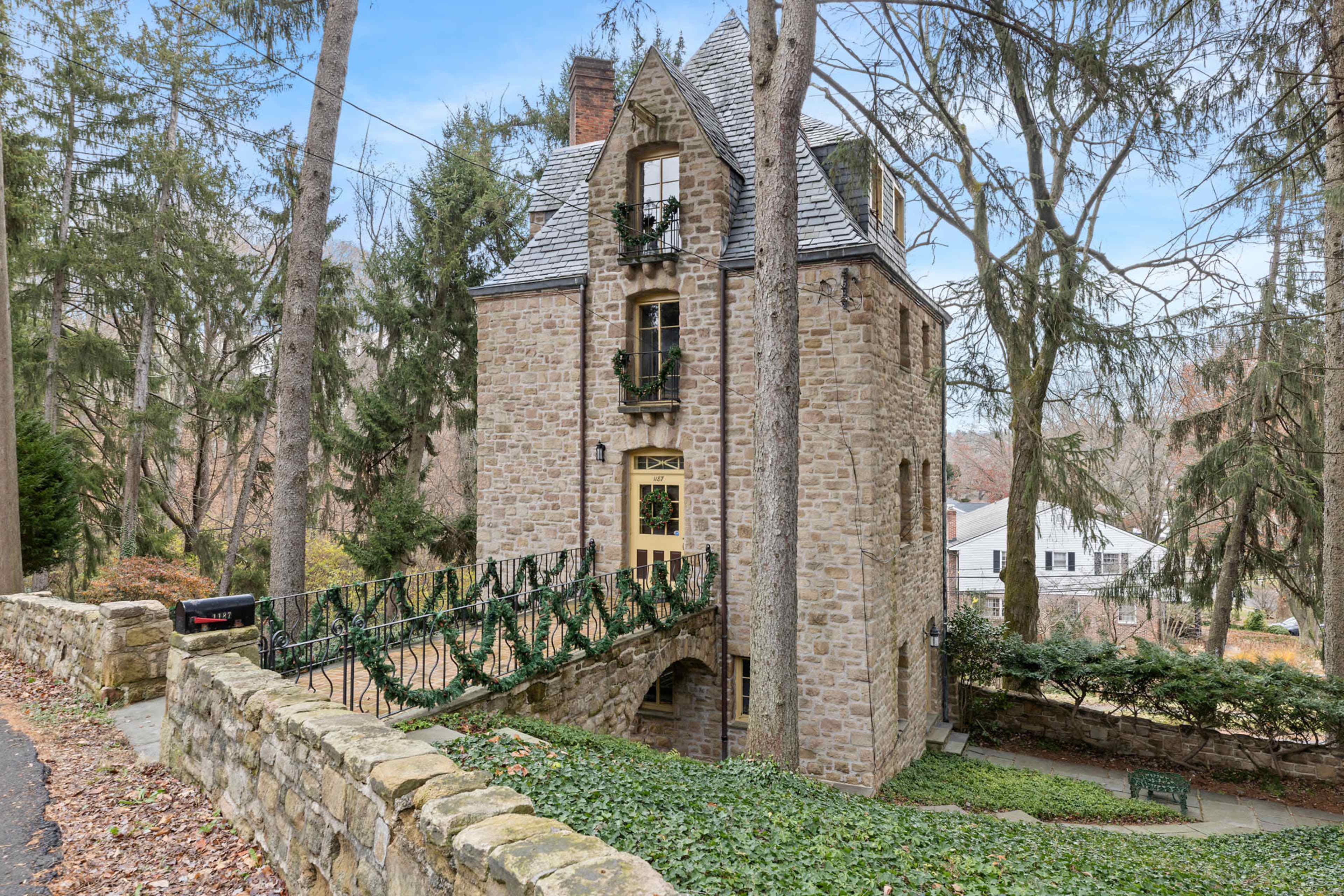 A tall stone house with a distinct architectural style is surrounded by trees and greenery along a pathway.