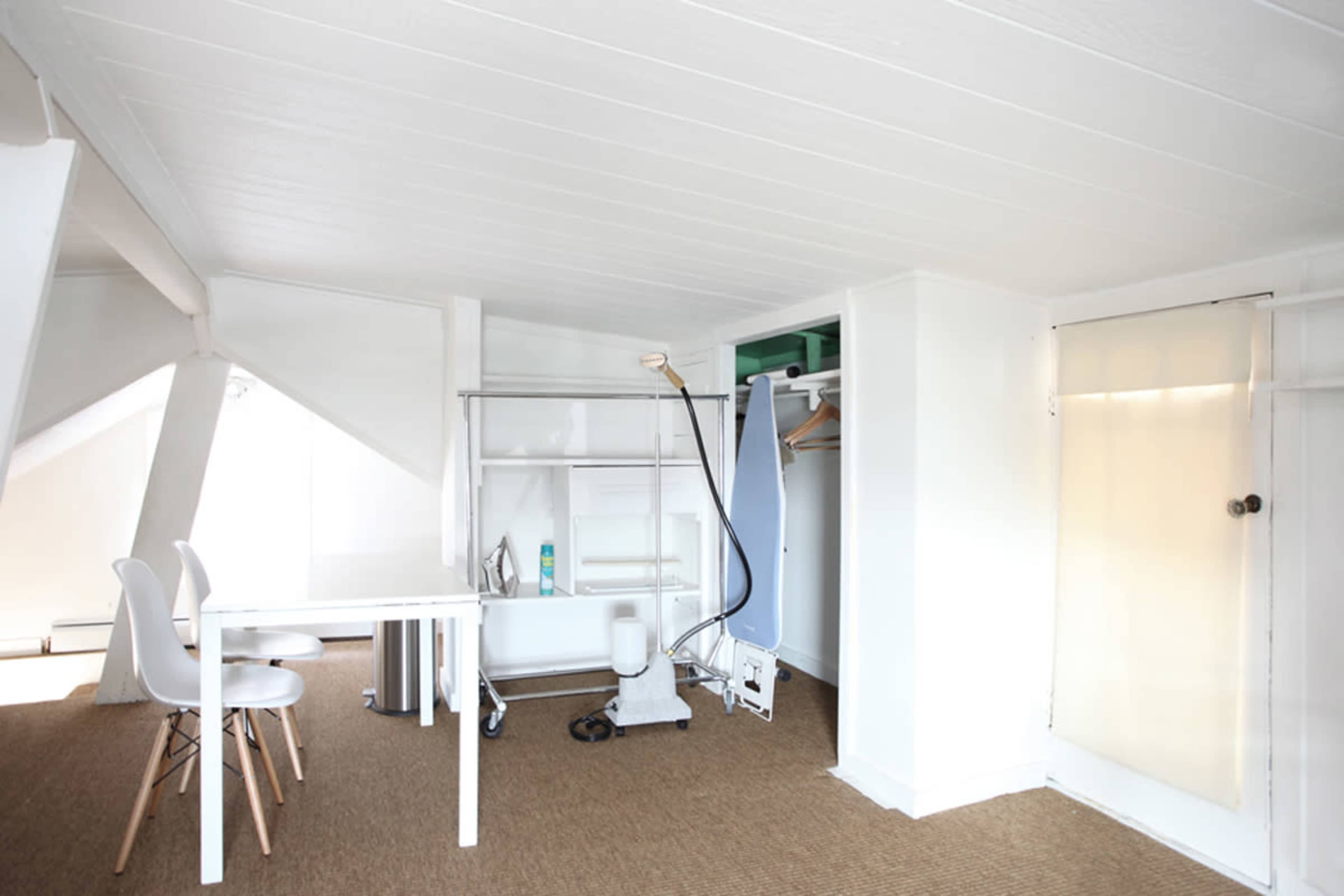 A bright, minimalist room featuring a small table, white chairs, a wardrobe, and an ironing board.