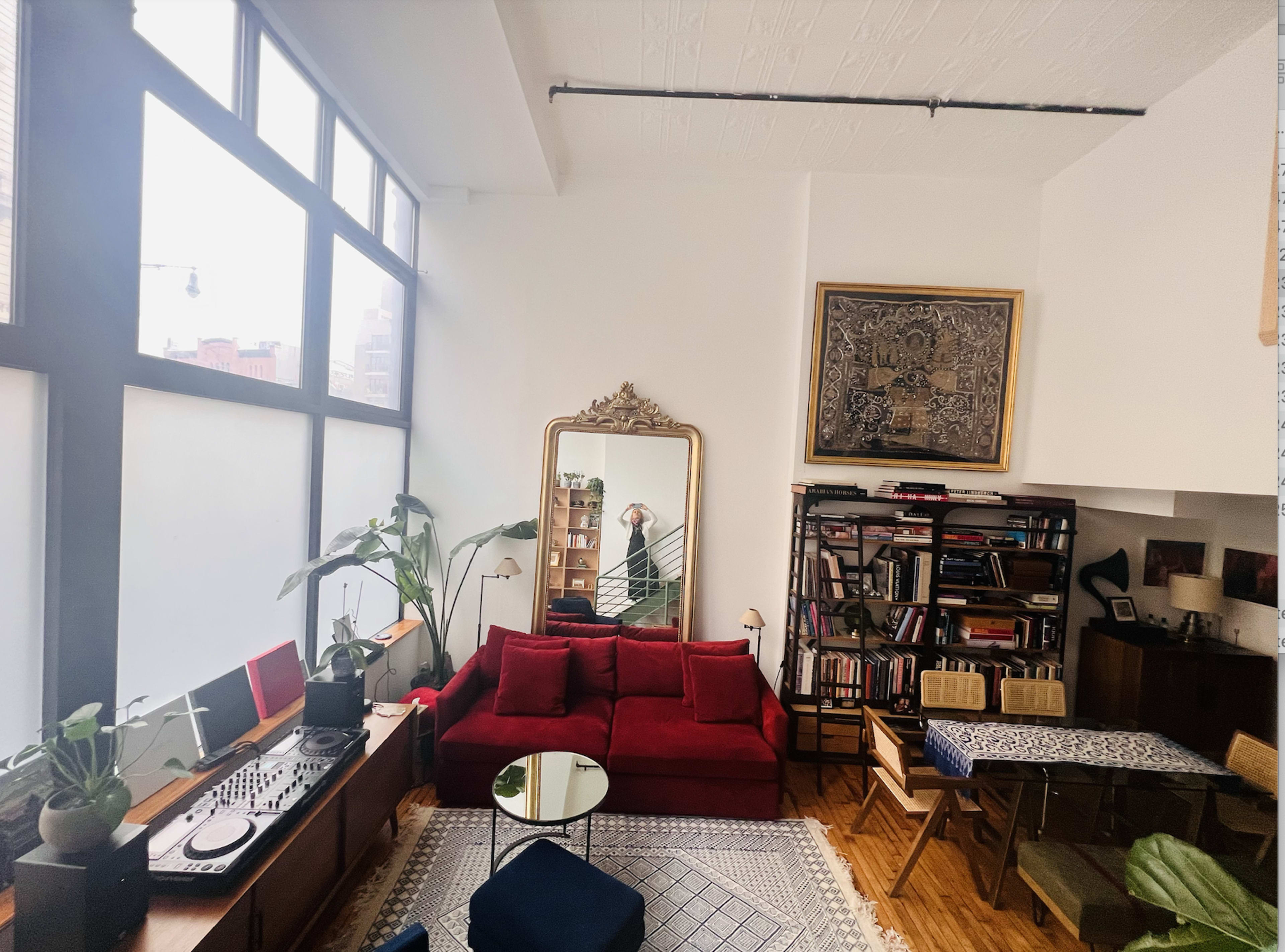 A bright living room featuring a red sofa, a large mirror, plants, bookshelves, and a dining table.