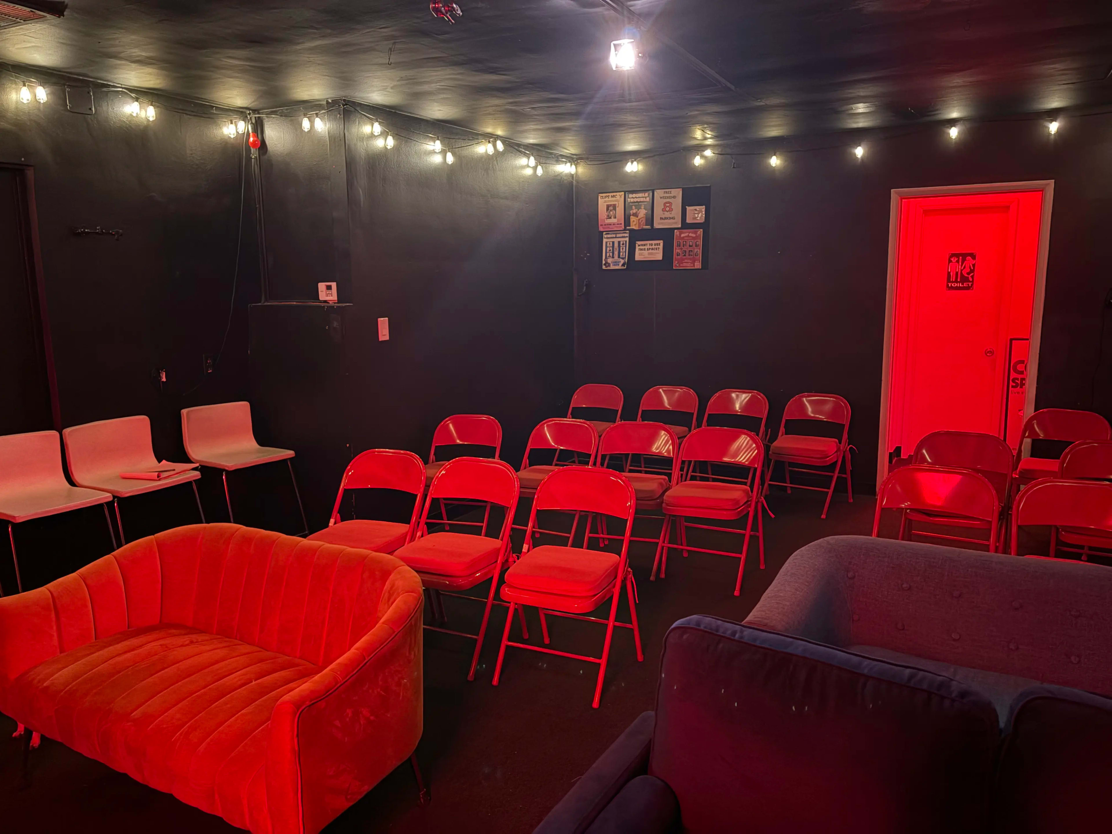 The image shows a small, dimly lit room with several red folding chairs arranged in two rows, a bright orange couch, and a door at the back.