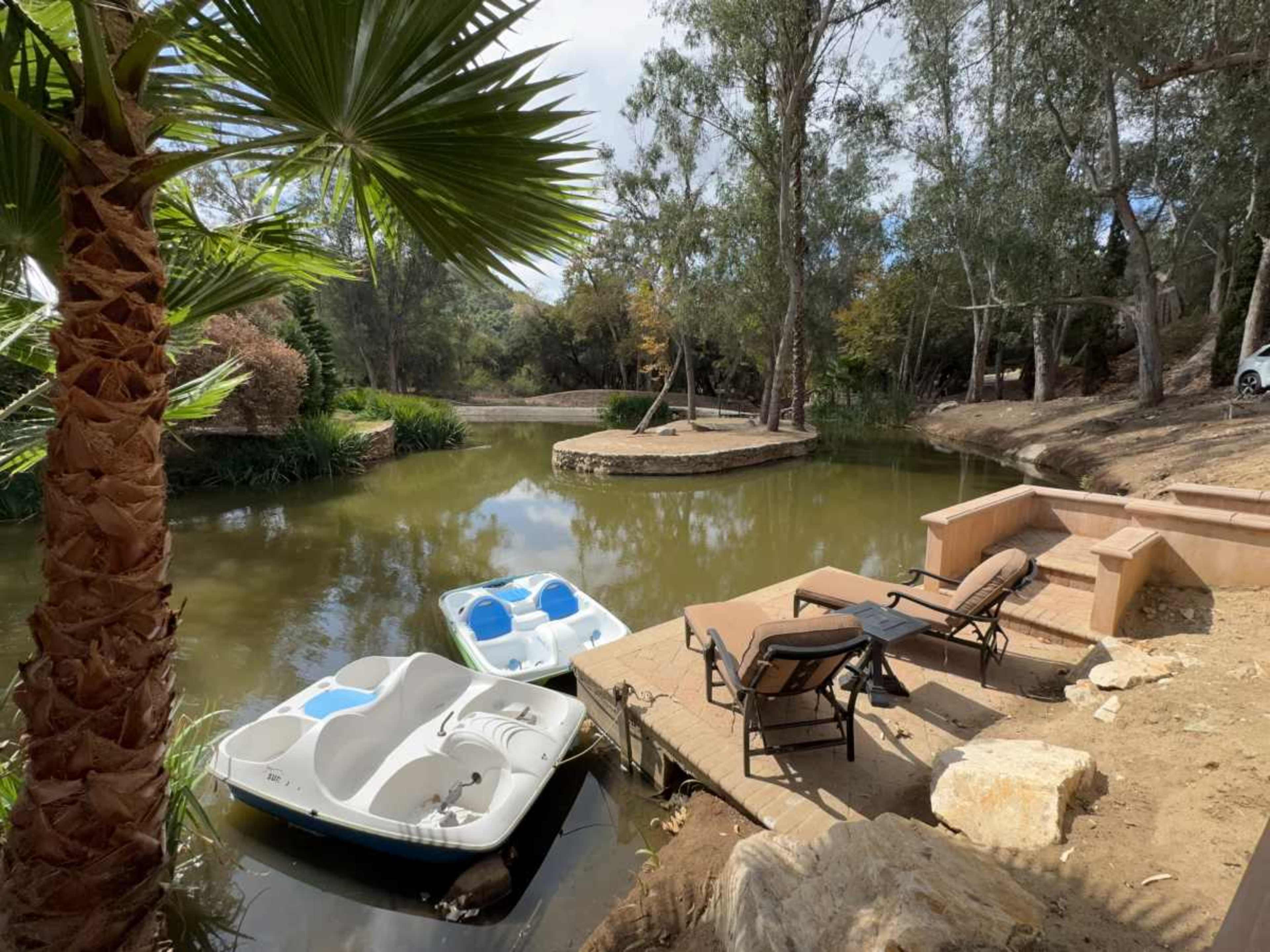 A calm riverbank scene features two boats moored near a wooden platform with lawn chairs and lush greenery surrounding the water.