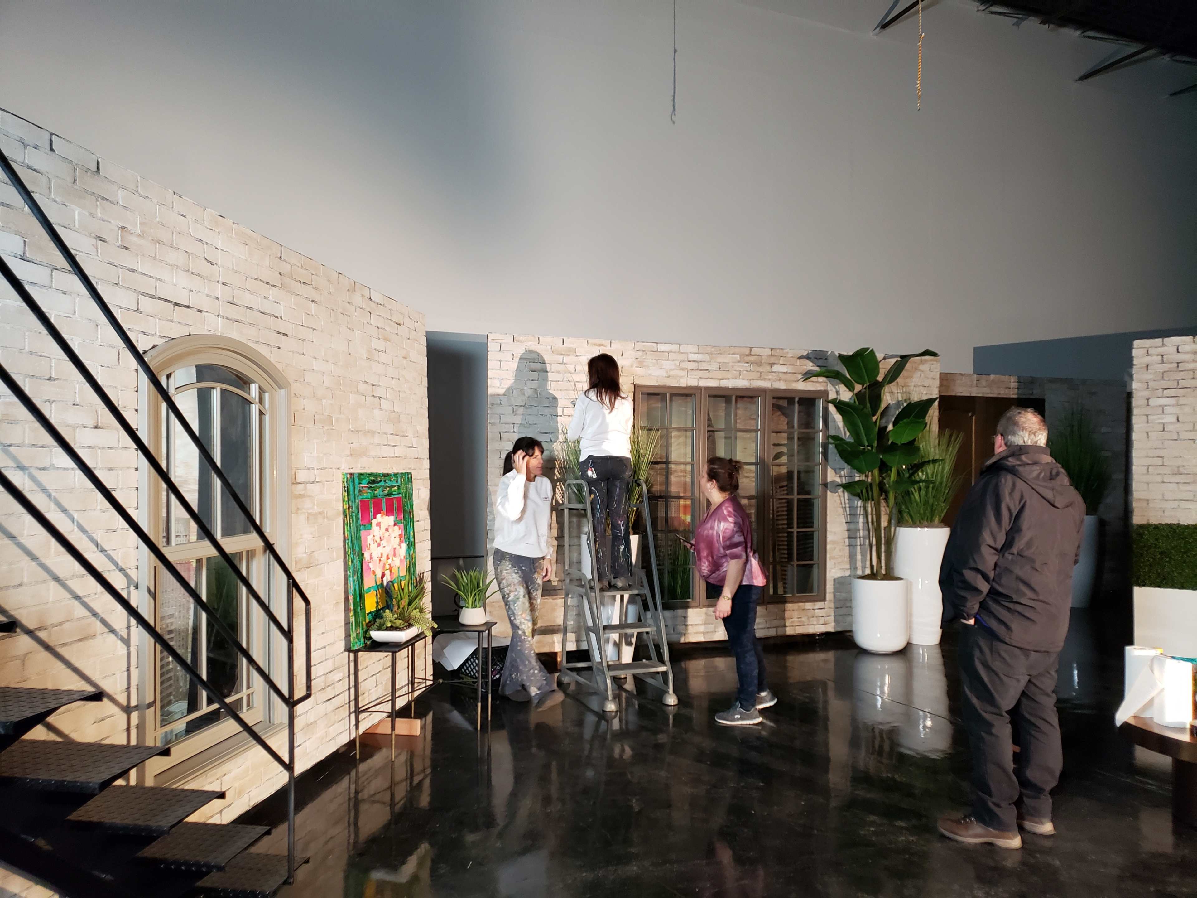 A group of four individuals are inside a spacious studio setting, with two people working on a ladder to adjust an object, while two others stand nearby observing.