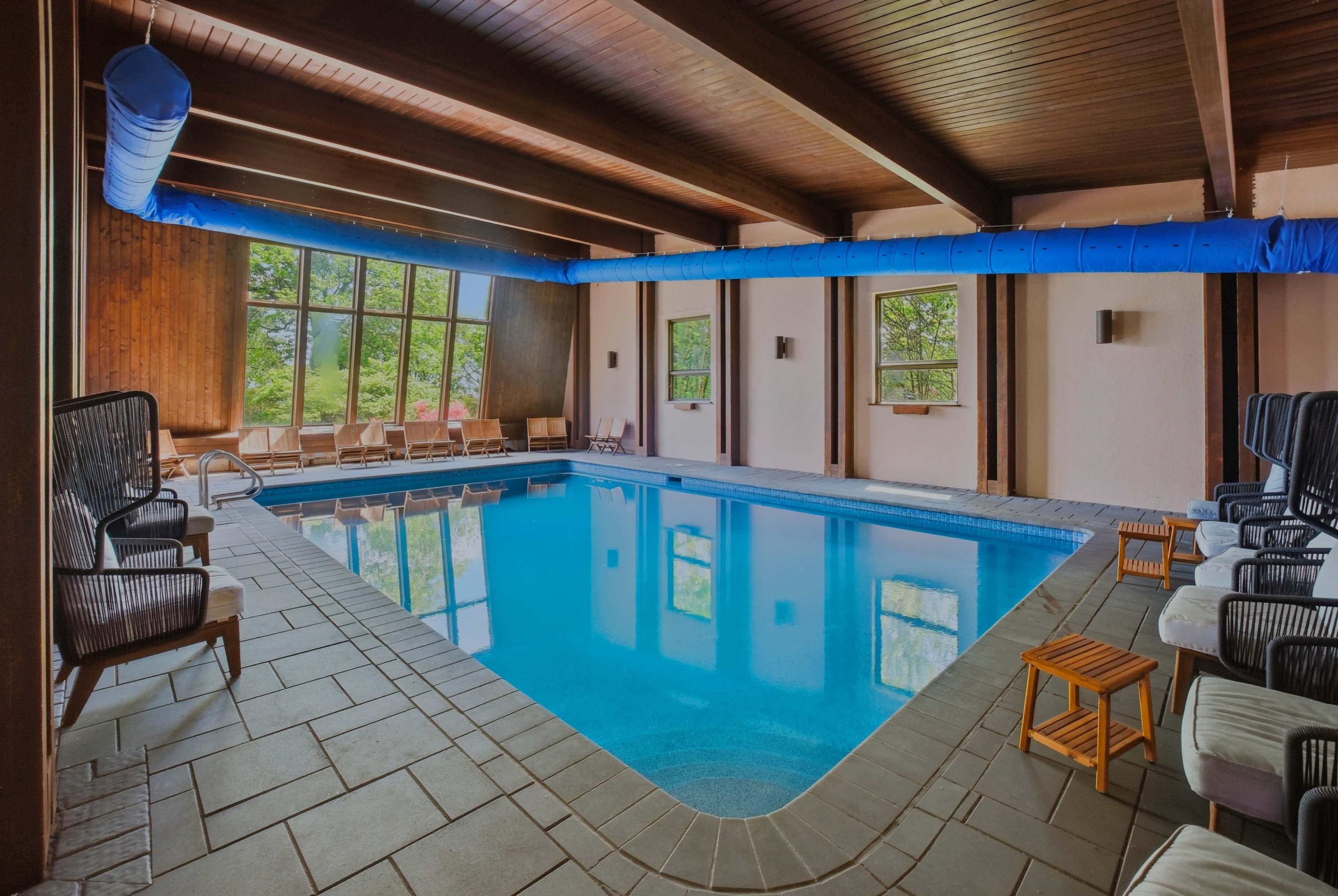 The image shows an indoor swimming pool surrounded by wooden beams and large windows that overlook a natural setting.