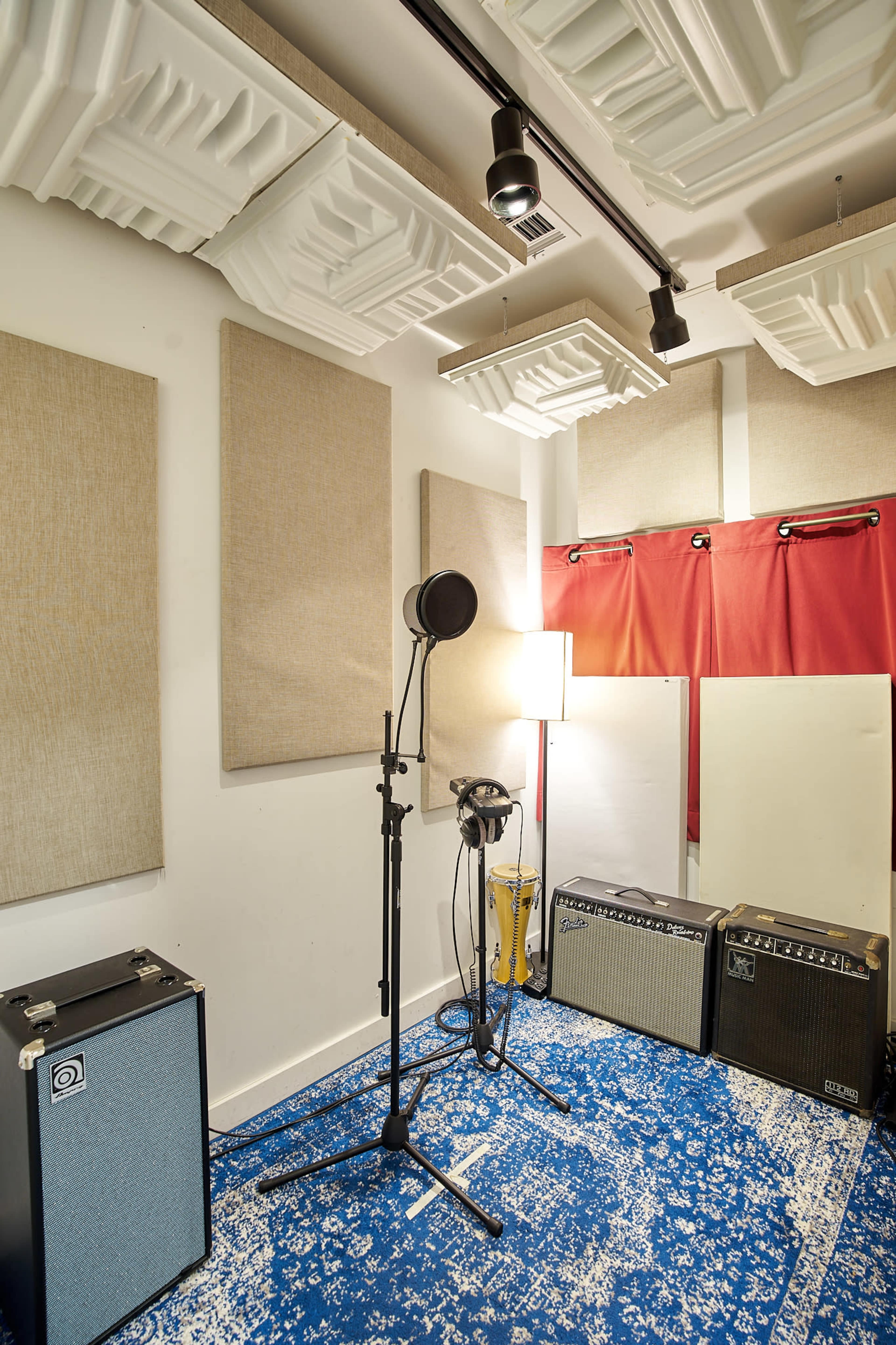 The image depicts a soundproof recording studio with acoustic panels on the walls, a microphone on a stand, a lamp, and various musical amplifiers.