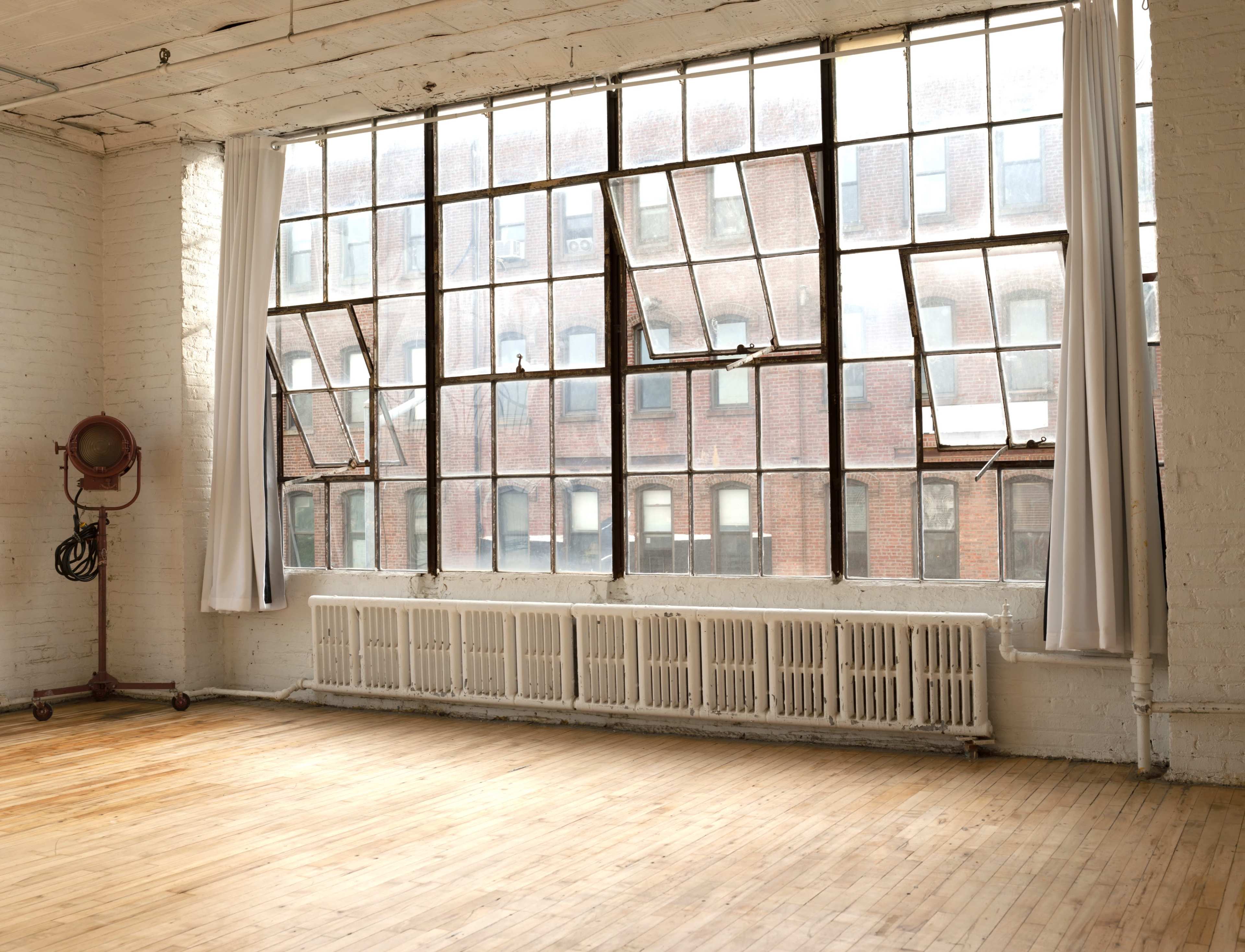 A spacious, light-filled room with large windows and wooden floors, featuring a radiator along the wall.