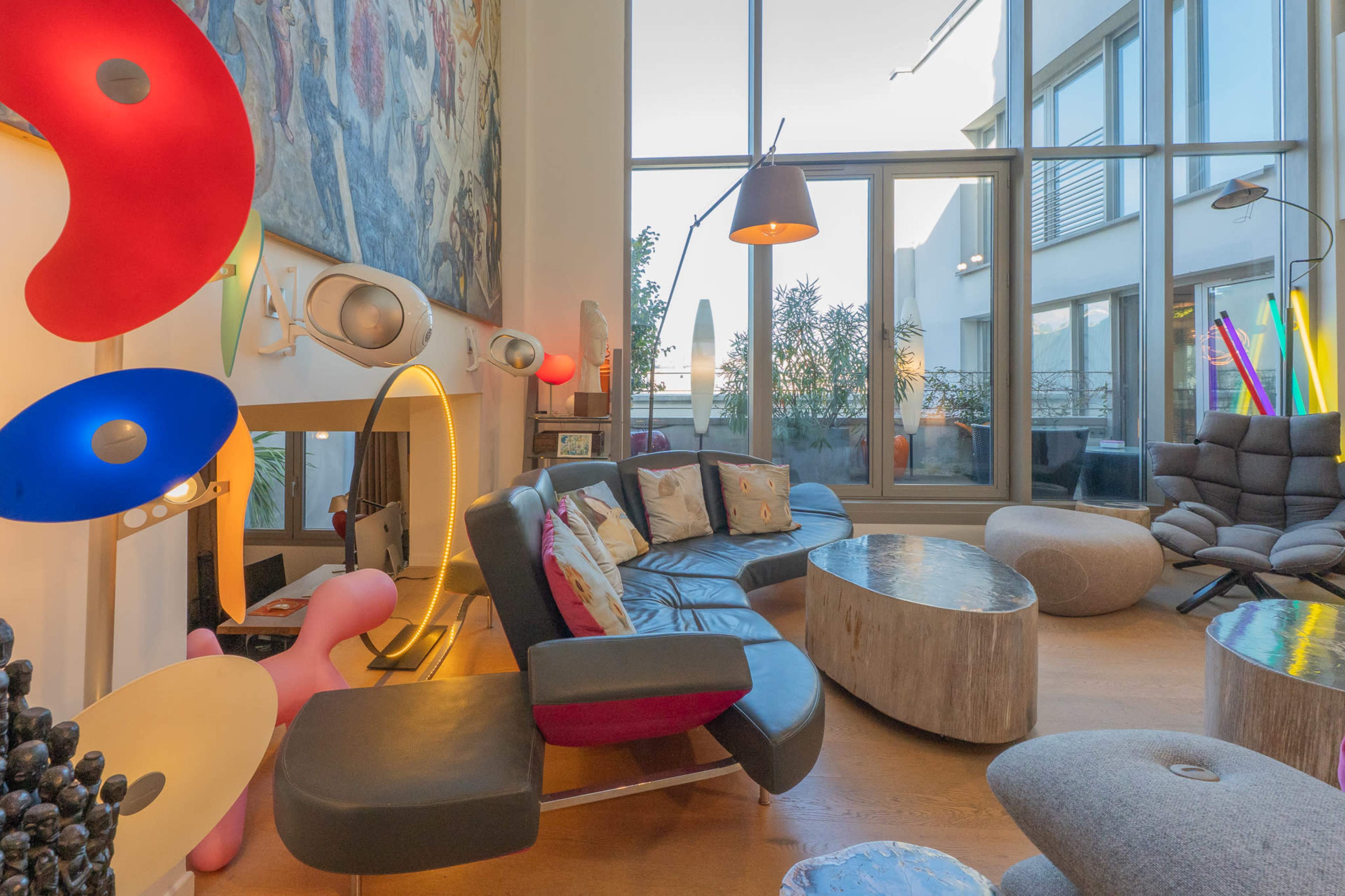 The image shows a modern living room featuring a variety of furniture styles, large windows, and colorful decorative elements.