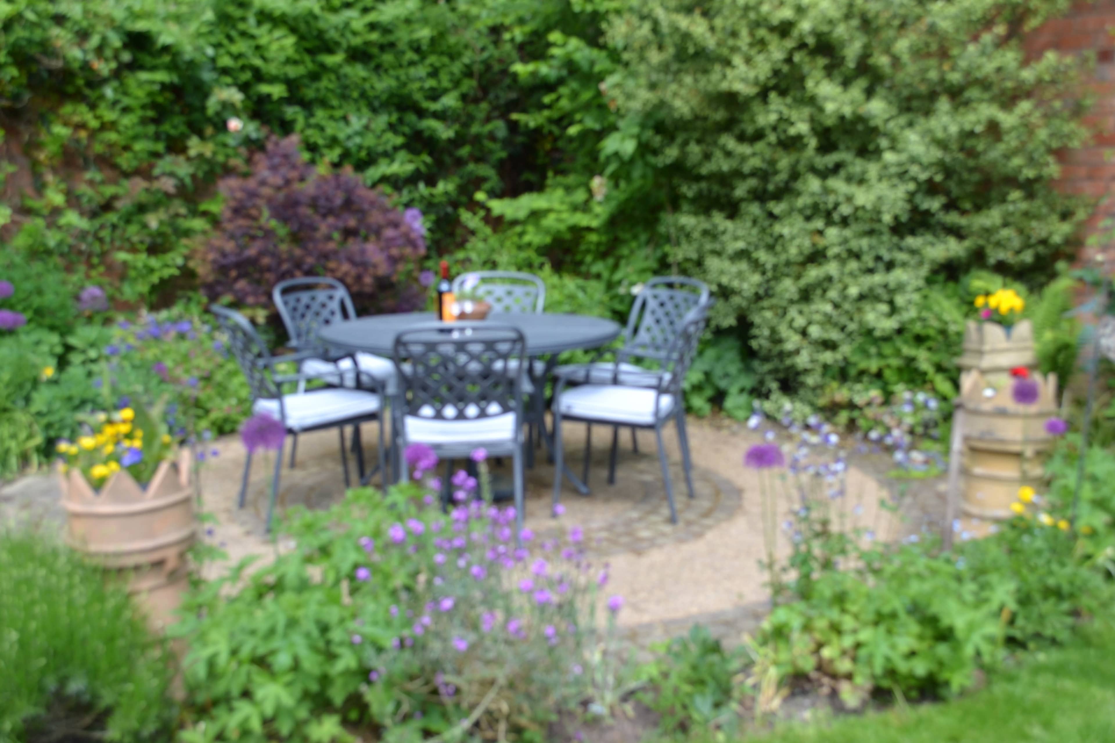 A circular table surrounded by four chairs sits on a stone path in a garden filled with various plants and flowers.