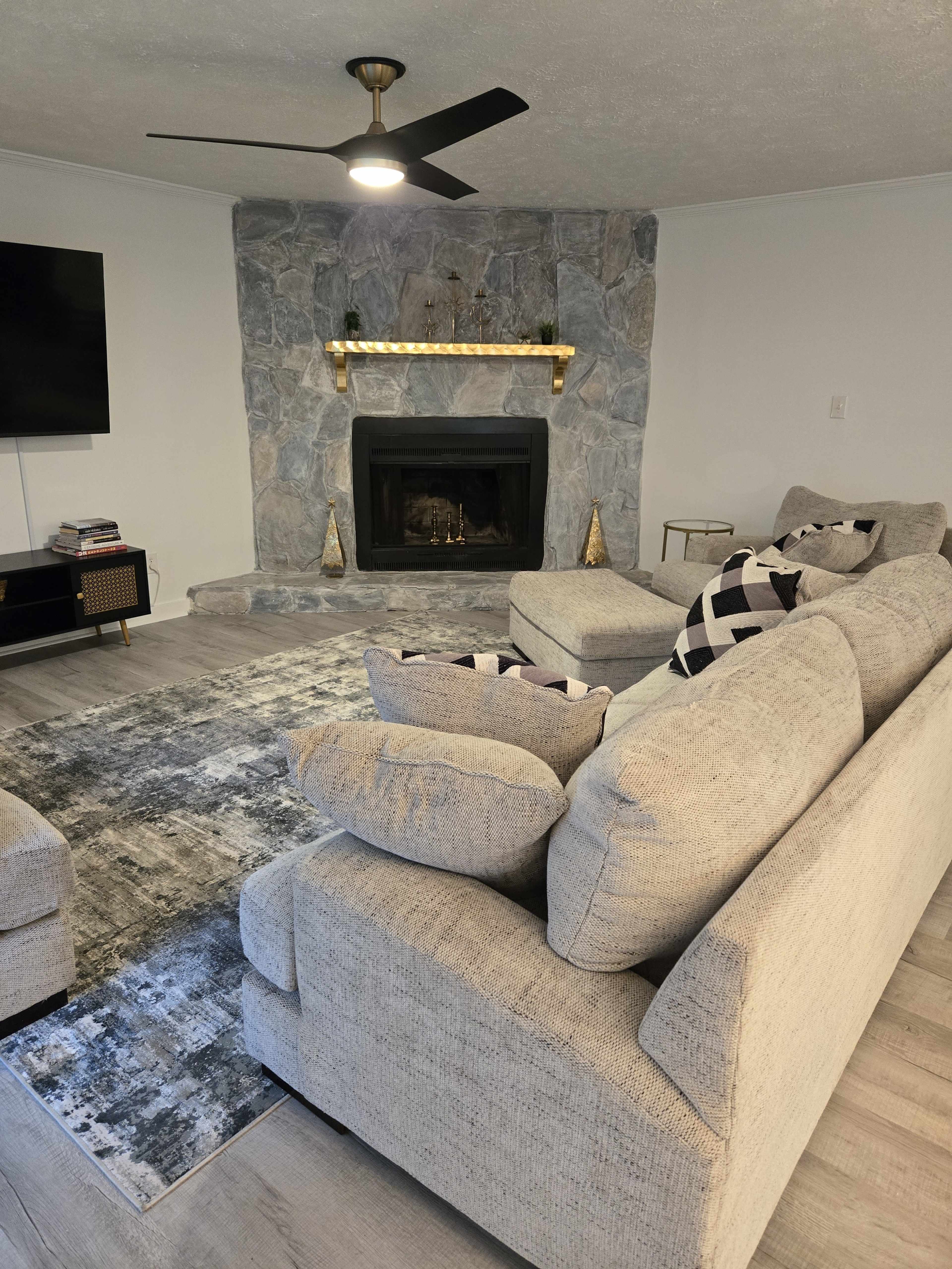 The image shows a modern living room with a stone fireplace, a large gray sectional sofa, and a decorative rug on a wooden floor.