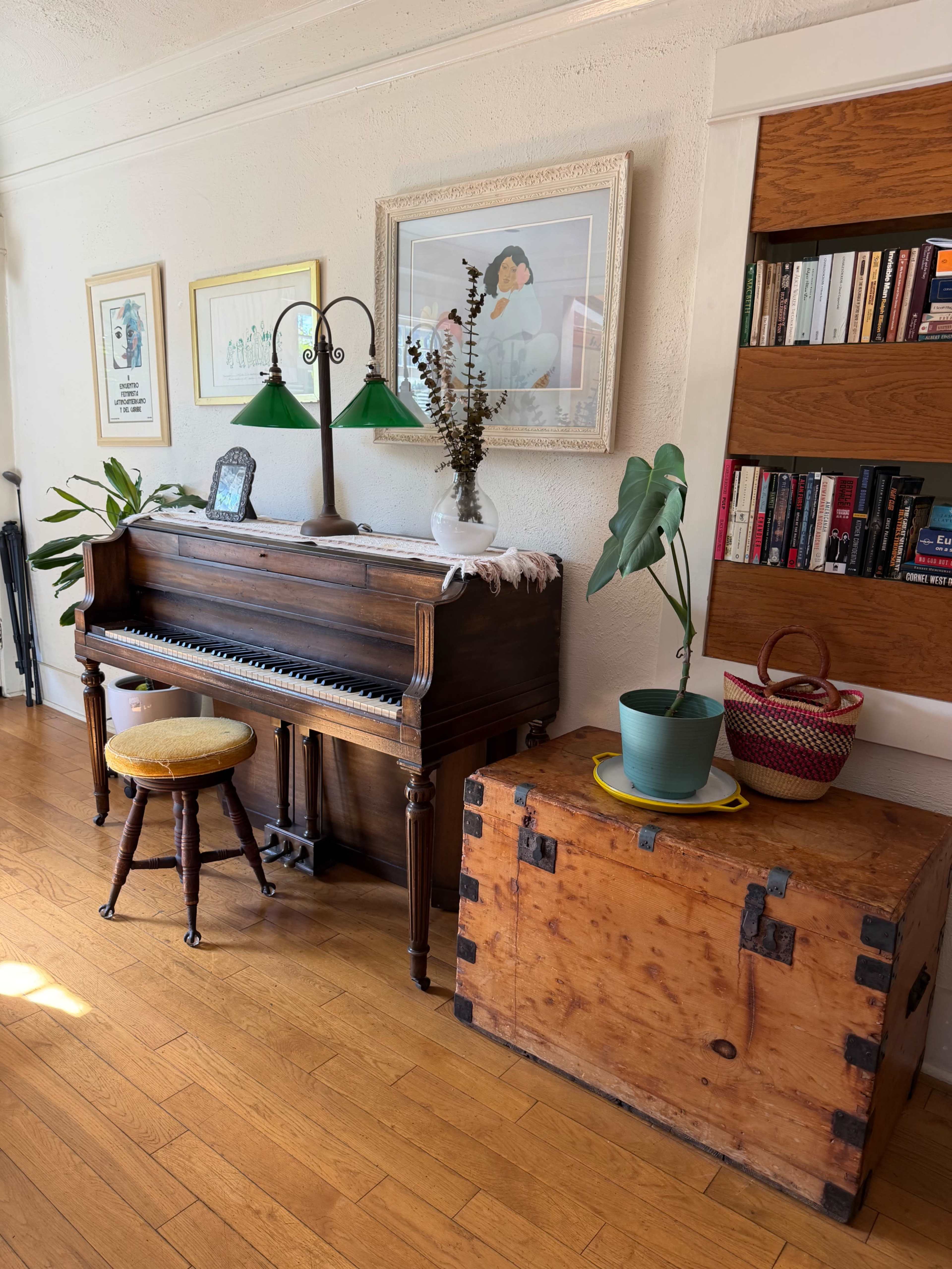 A vintage piano is placed against a wall, accompanied by a green lamp, a houseplant, and a wooden trunk, while framed artworks hang above.