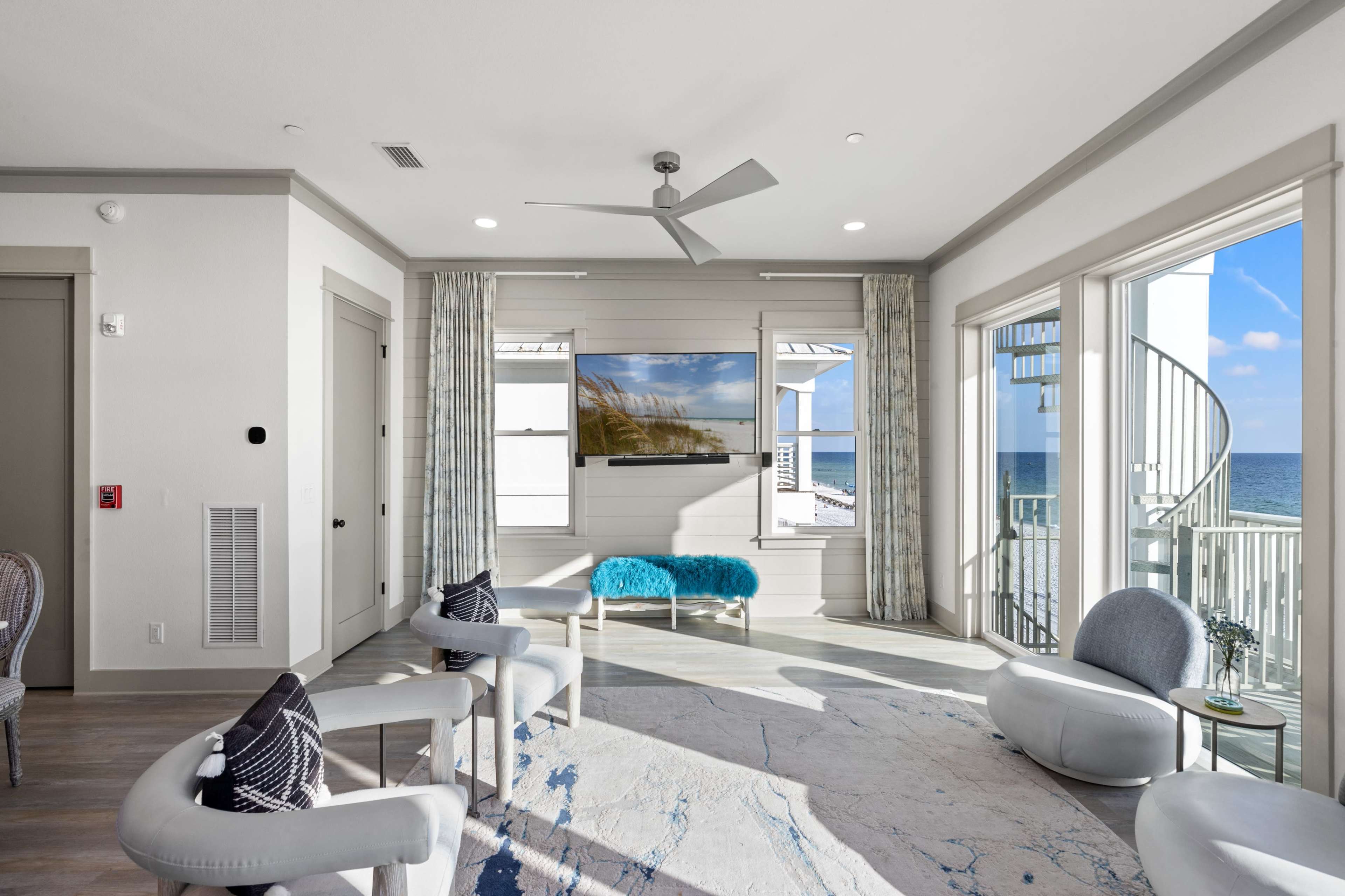 A bright and airy living room features a large window with ocean views, a mounted television, and modern furniture arranged around a light-colored rug.