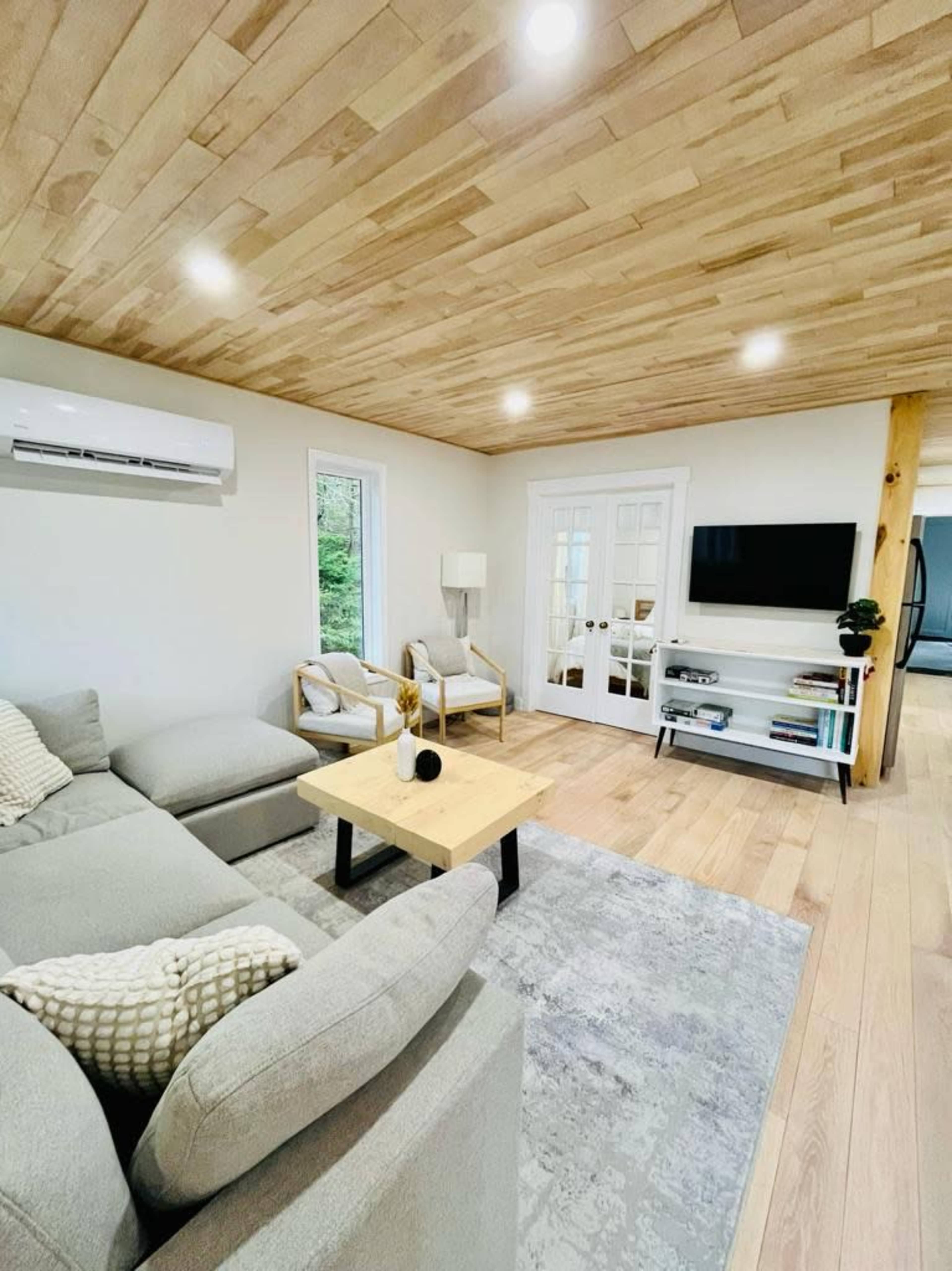 The image shows a modern living room with a light wooden ceiling, a gray sectional sofa, a coffee table, and a television mounted on the wall opposite a set of glass-paned doors.