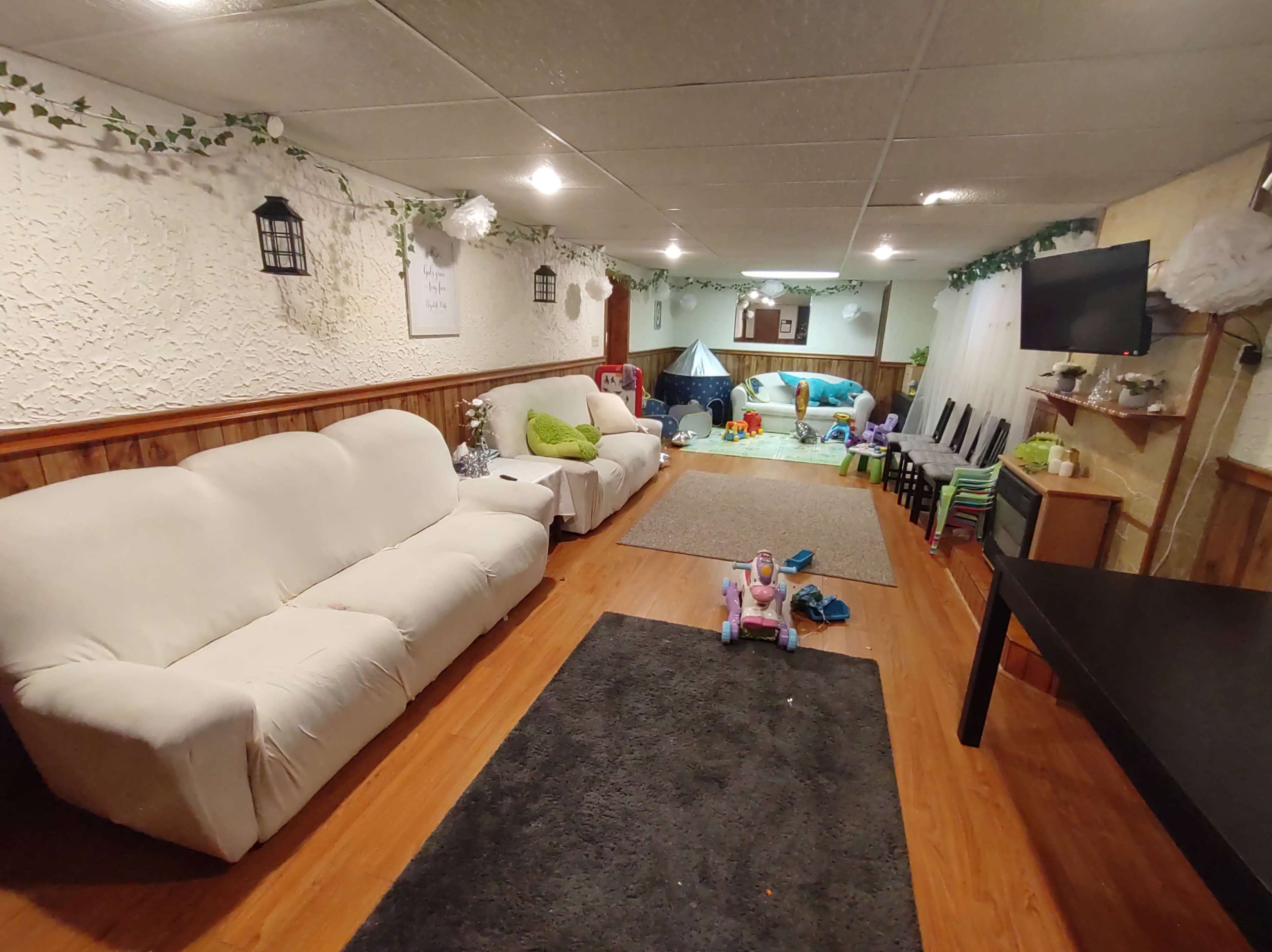 A spacious basement area furnished with white sofas, a black table, and various children's toys scattered across the floor.