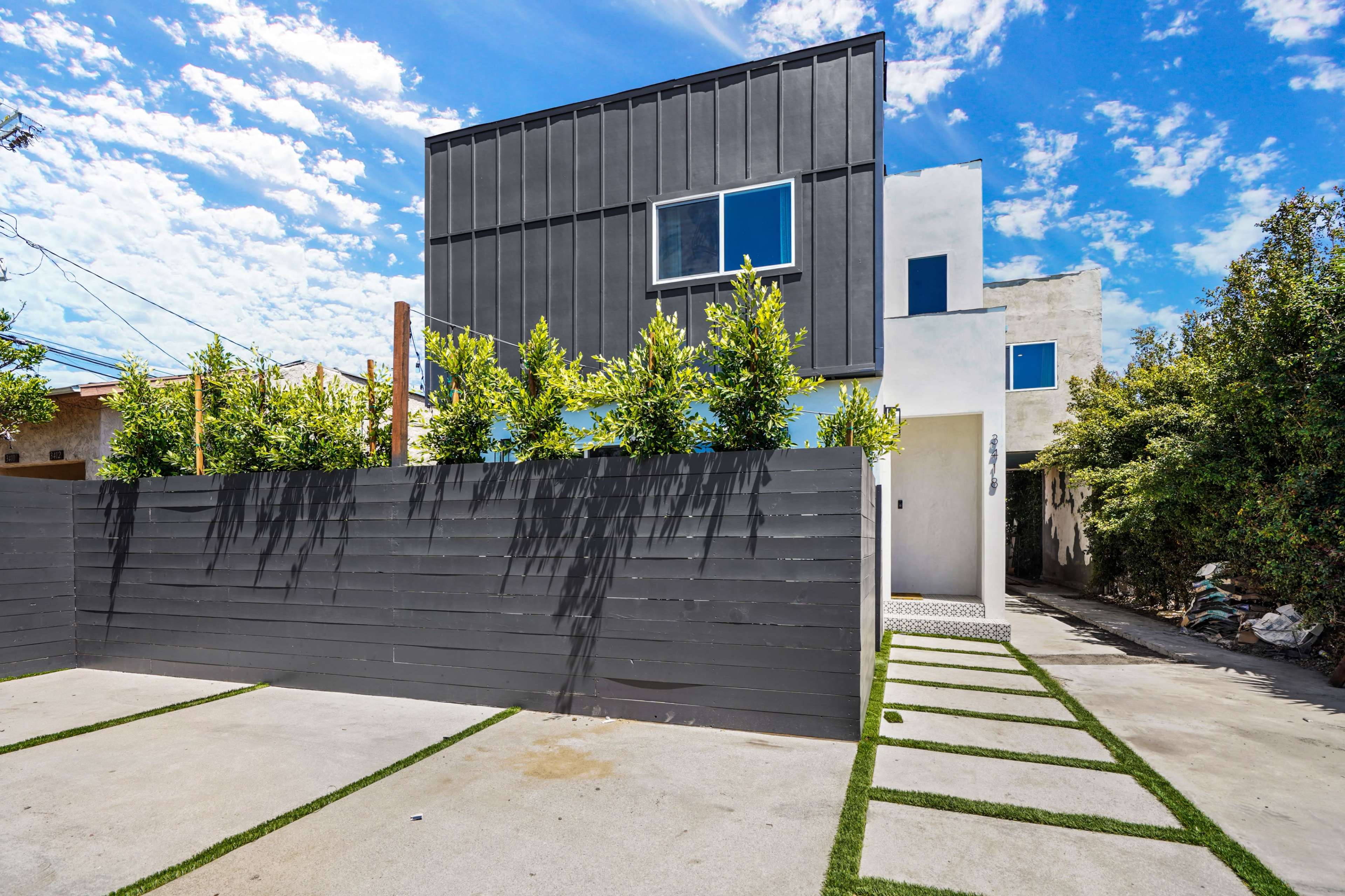 A modern two-story building with a black metal exterior is situated behind a gray fence, with neatly arranged concrete pathways and landscaped greenery in the front.