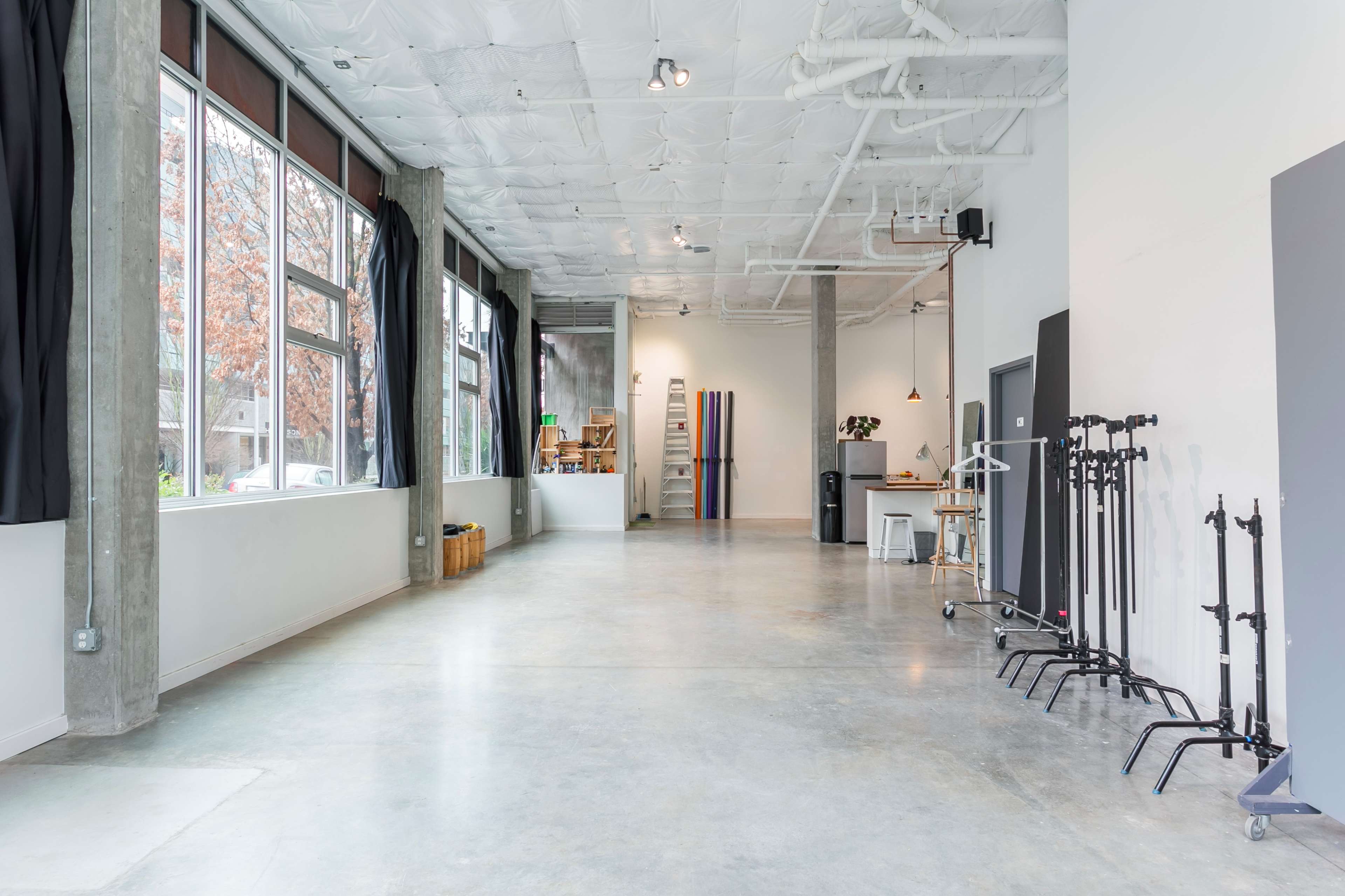 The image shows a spacious, well-lit studio with concrete floors, large windows, and various equipment organized along one side.