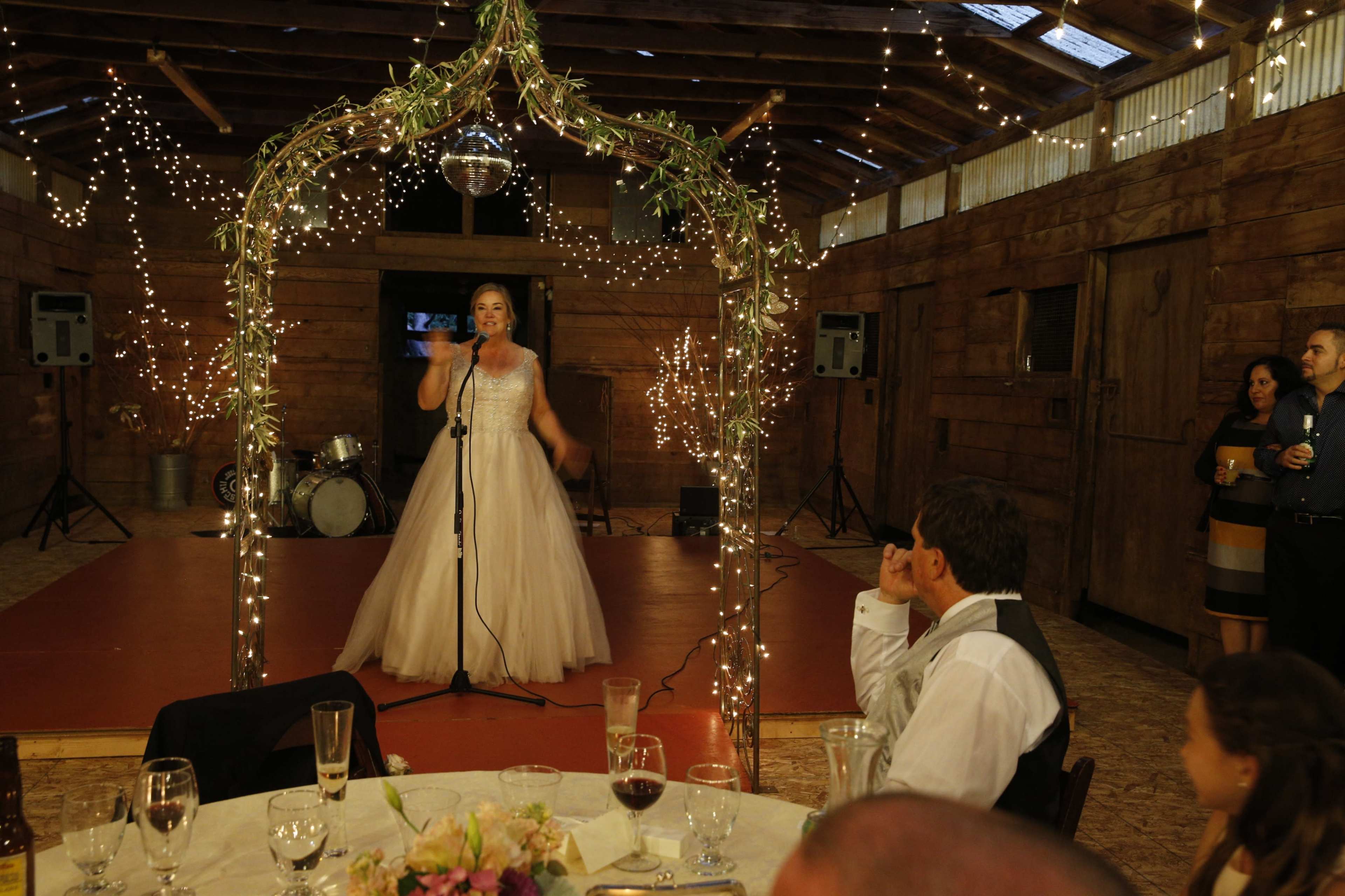 A woman in a formal dress stands on a small stage, singing into a microphone, while guests seated at tables observe in a rustic venue decorated with string lights.