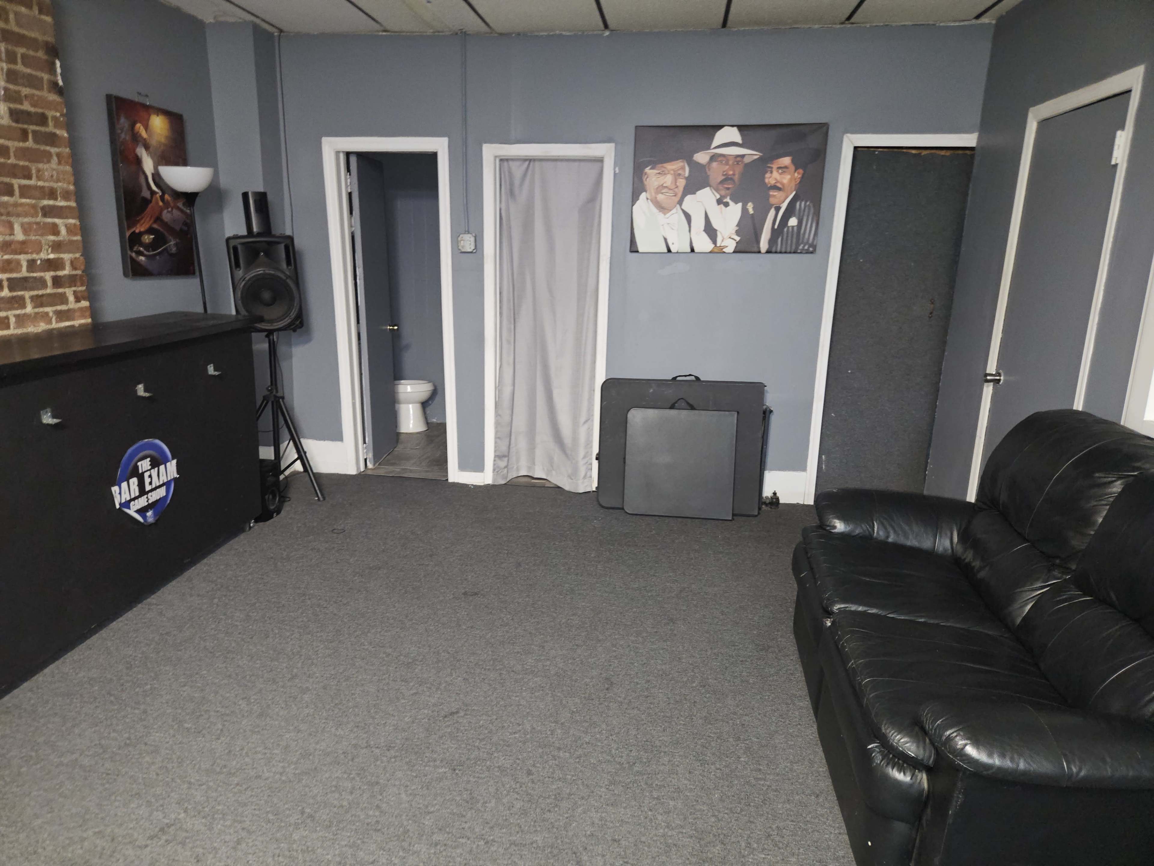 The image shows a sparsely furnished room with a black leather couch, a speaker near the wall, and a doorway leading to a bathroom in the background.