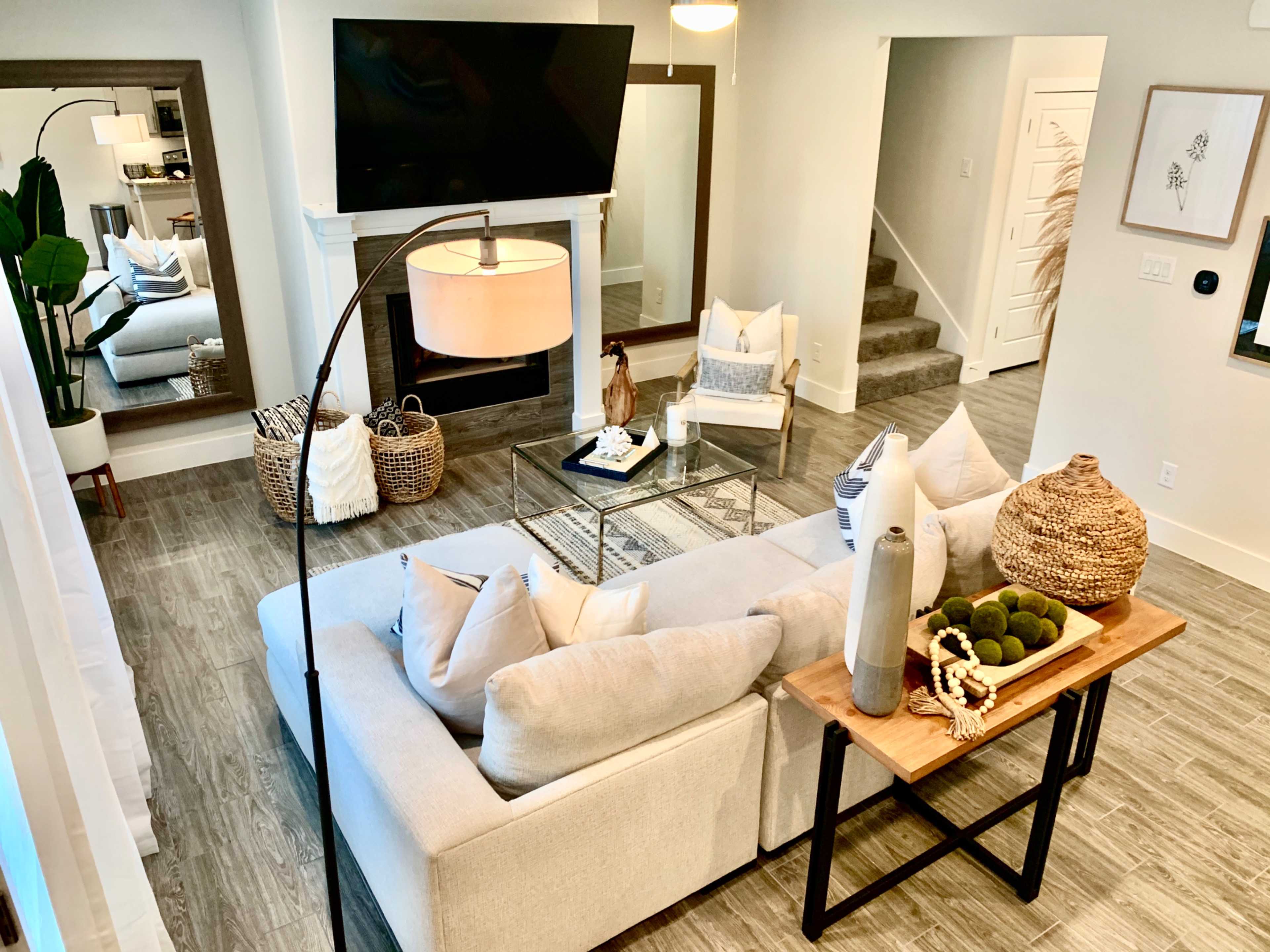 A modern living room featuring a light-colored sectional sofa, a glass coffee table, and a large television mounted above a fireplace, with decorative elements and a staircase visible in the background.