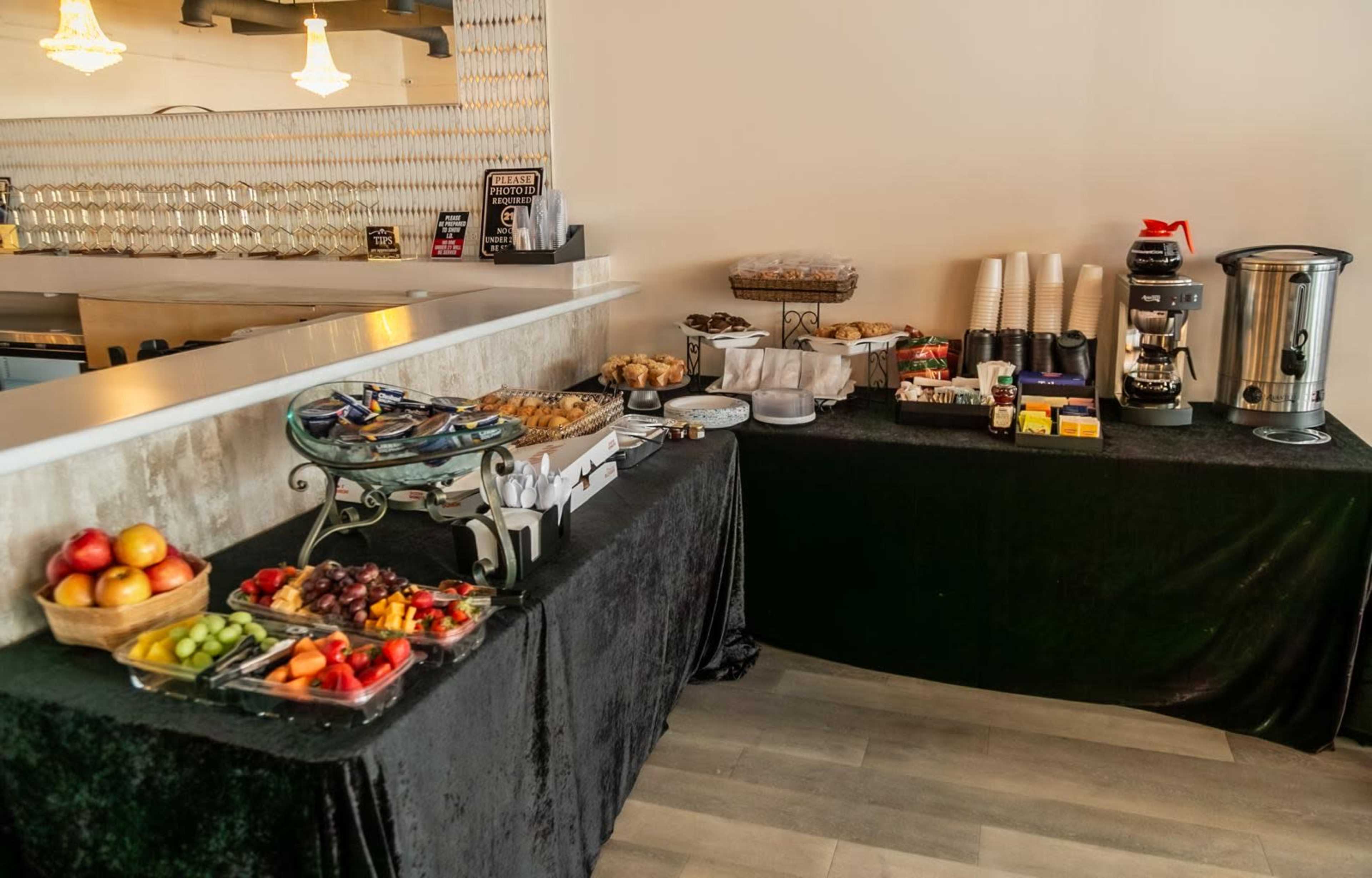 The image shows a buffet table set with a variety of fruits, pastries, coffee, and cups, arranged on a black tablecloth near a bar area.