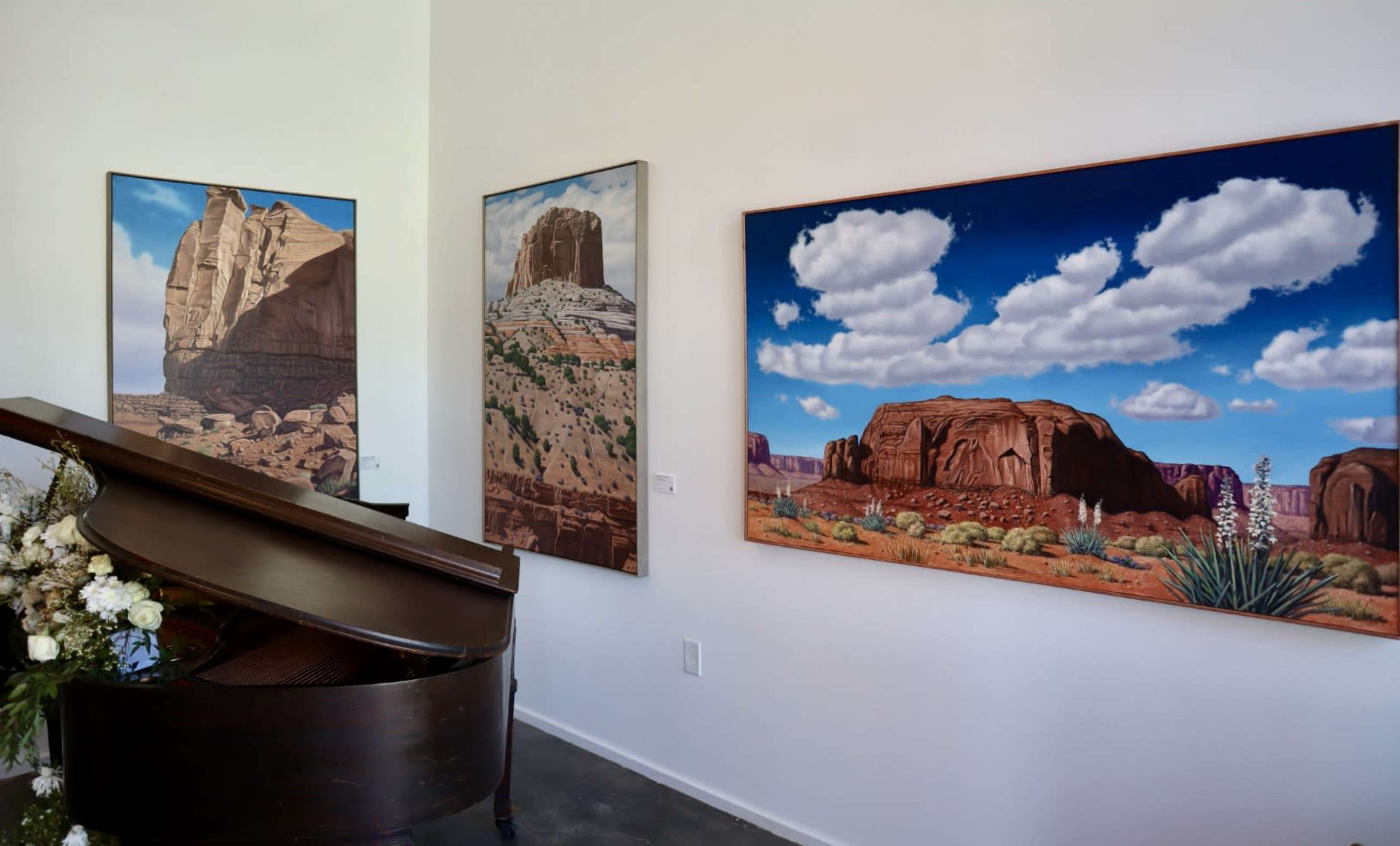 A grand piano sits in the foreground, while two landscape paintings featuring desert scenes are displayed on the white walls behind it.