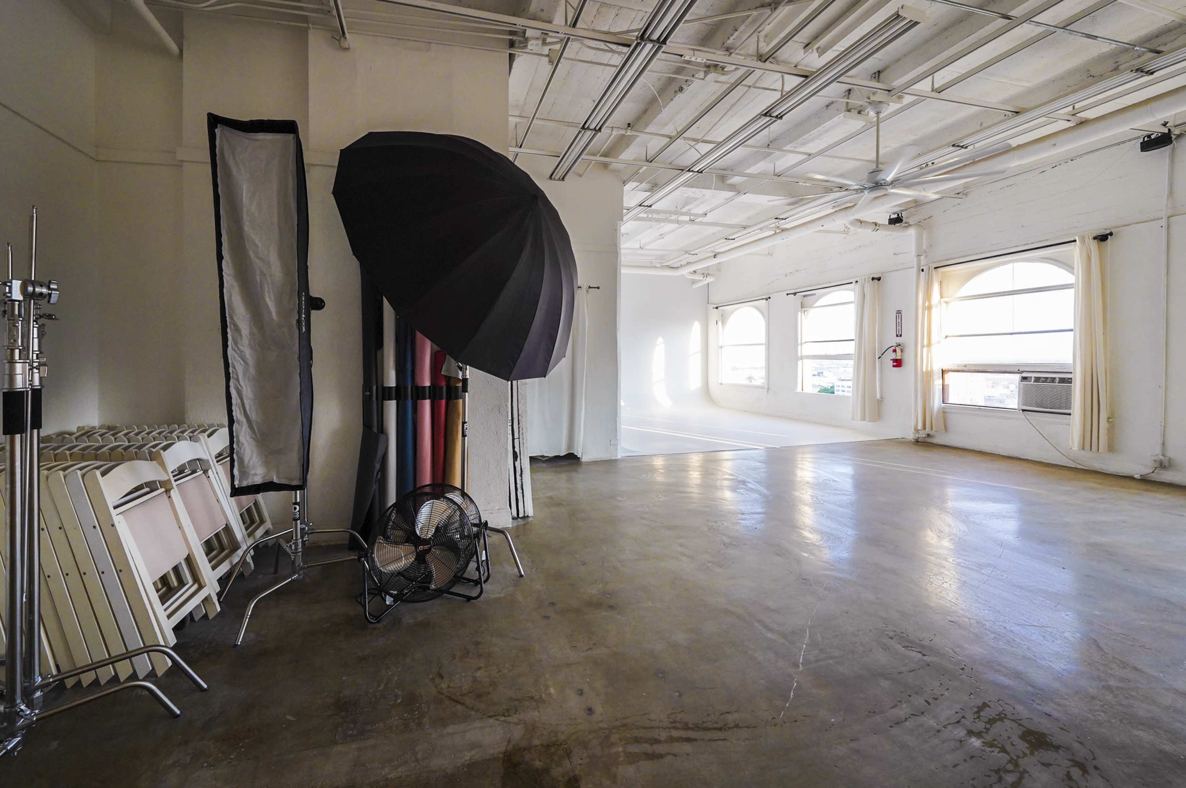 The image shows a spacious photography studio with a large umbrella light, folding chairs, and a fan against a wall, featuring large windows that allow natural light to enter.