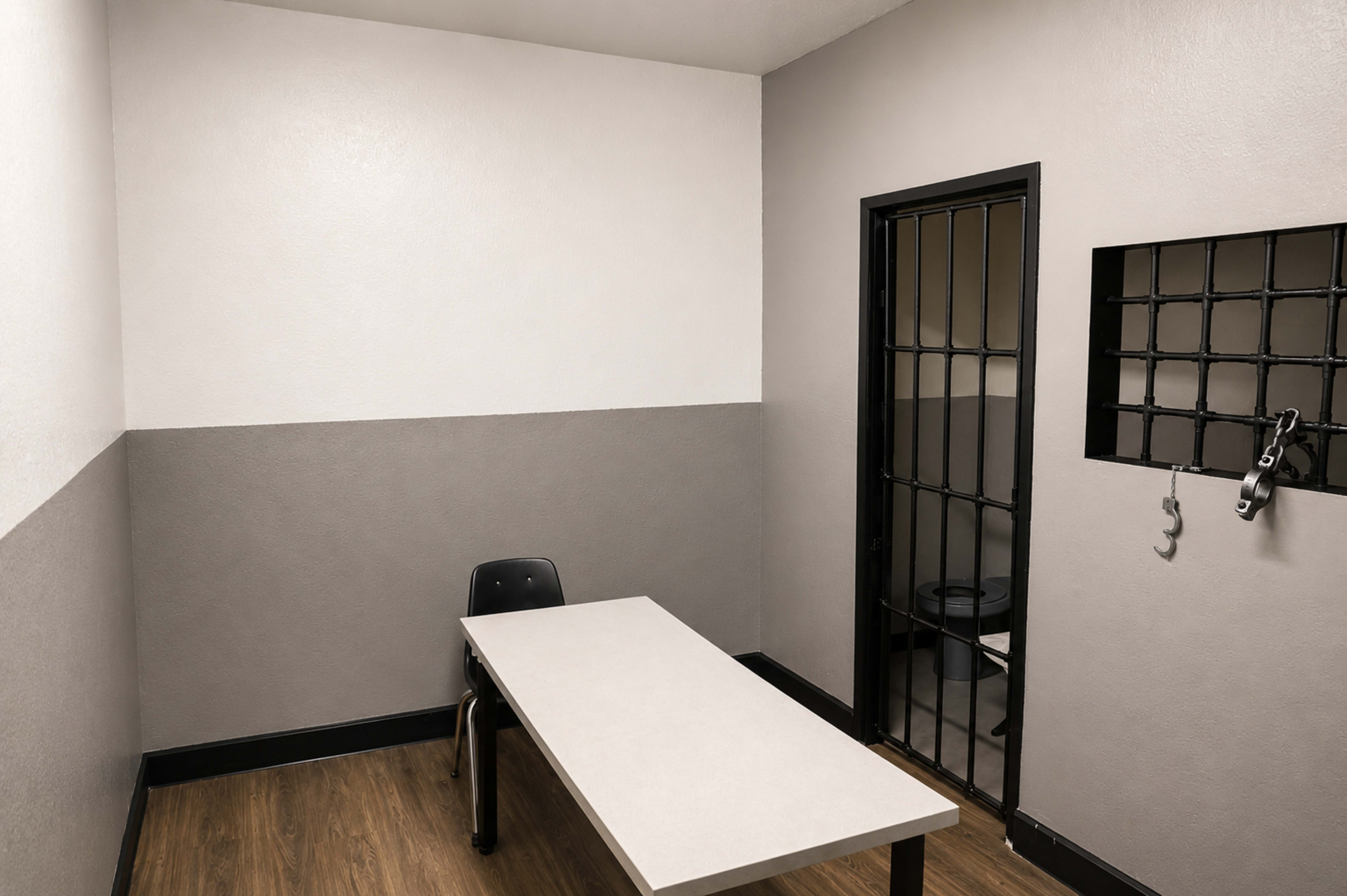 The image shows a small, empty interrogation room featuring a table and a black chair, with a barred door and a window displaying a toilet.