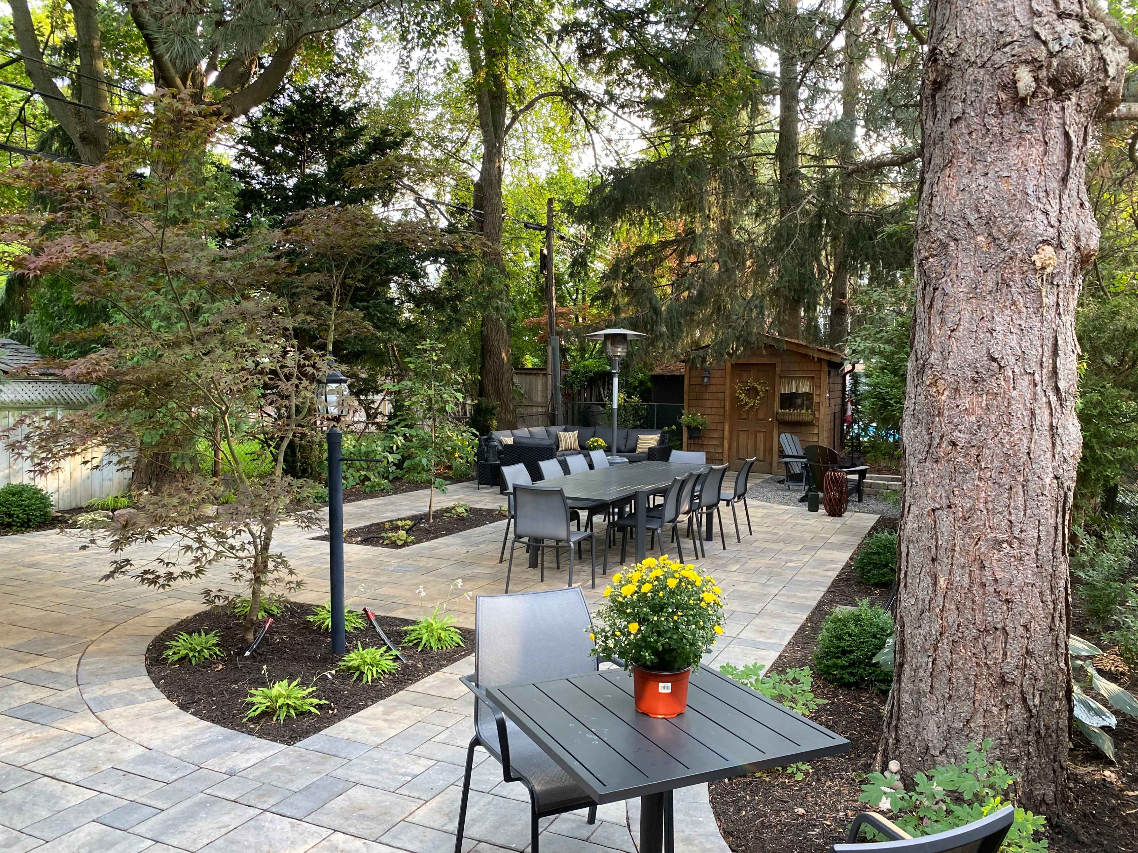 The image shows a landscaped patio area with a long table surrounded by chairs, a small potted plant, and a wooden shed in the background.