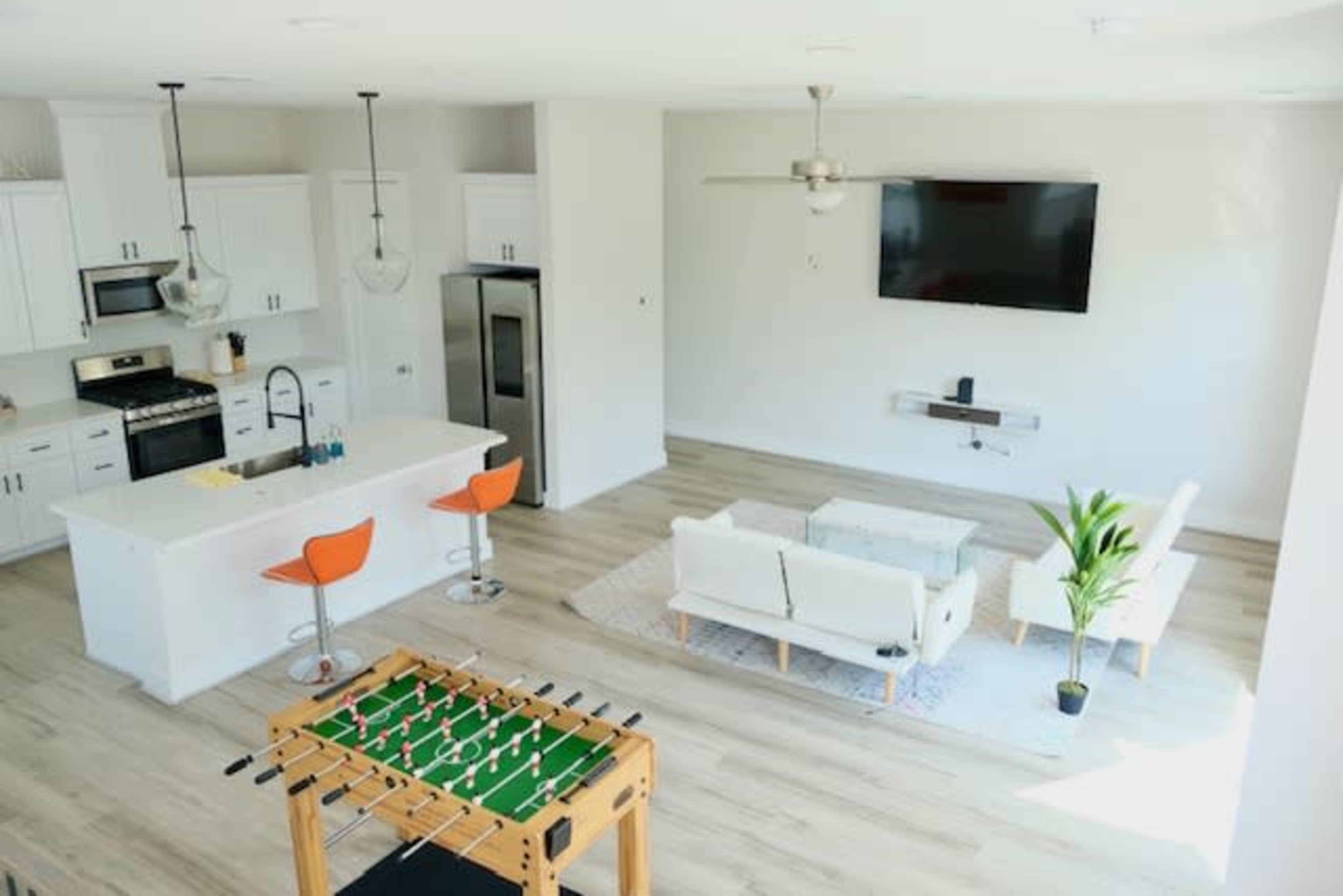 A modern living space showcases a kitchen with bar stools, a foosball table, a sofa set, and a wall-mounted television.