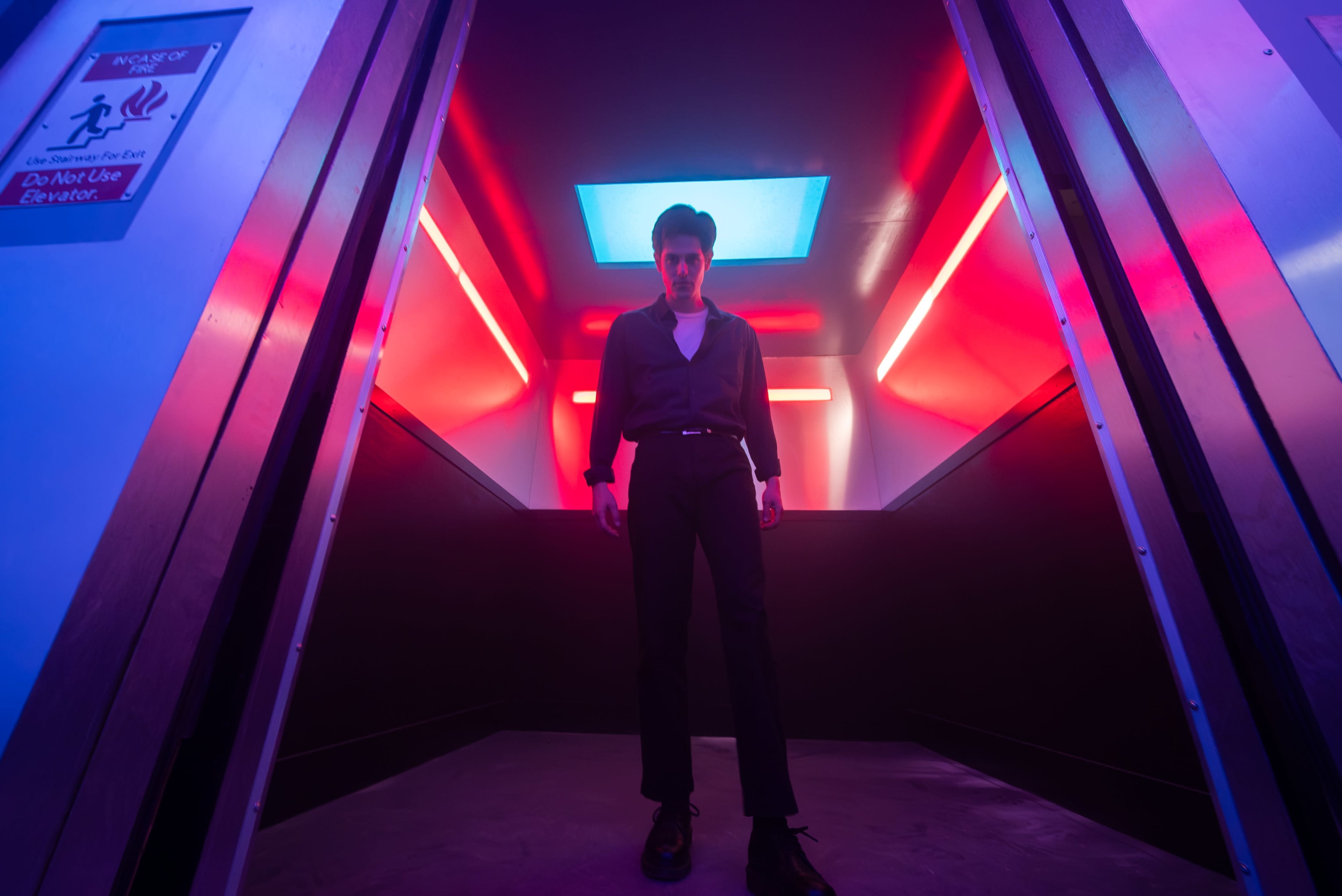 A figure stands in a dimly lit elevator with vibrant blue and red lights illuminating the space.
