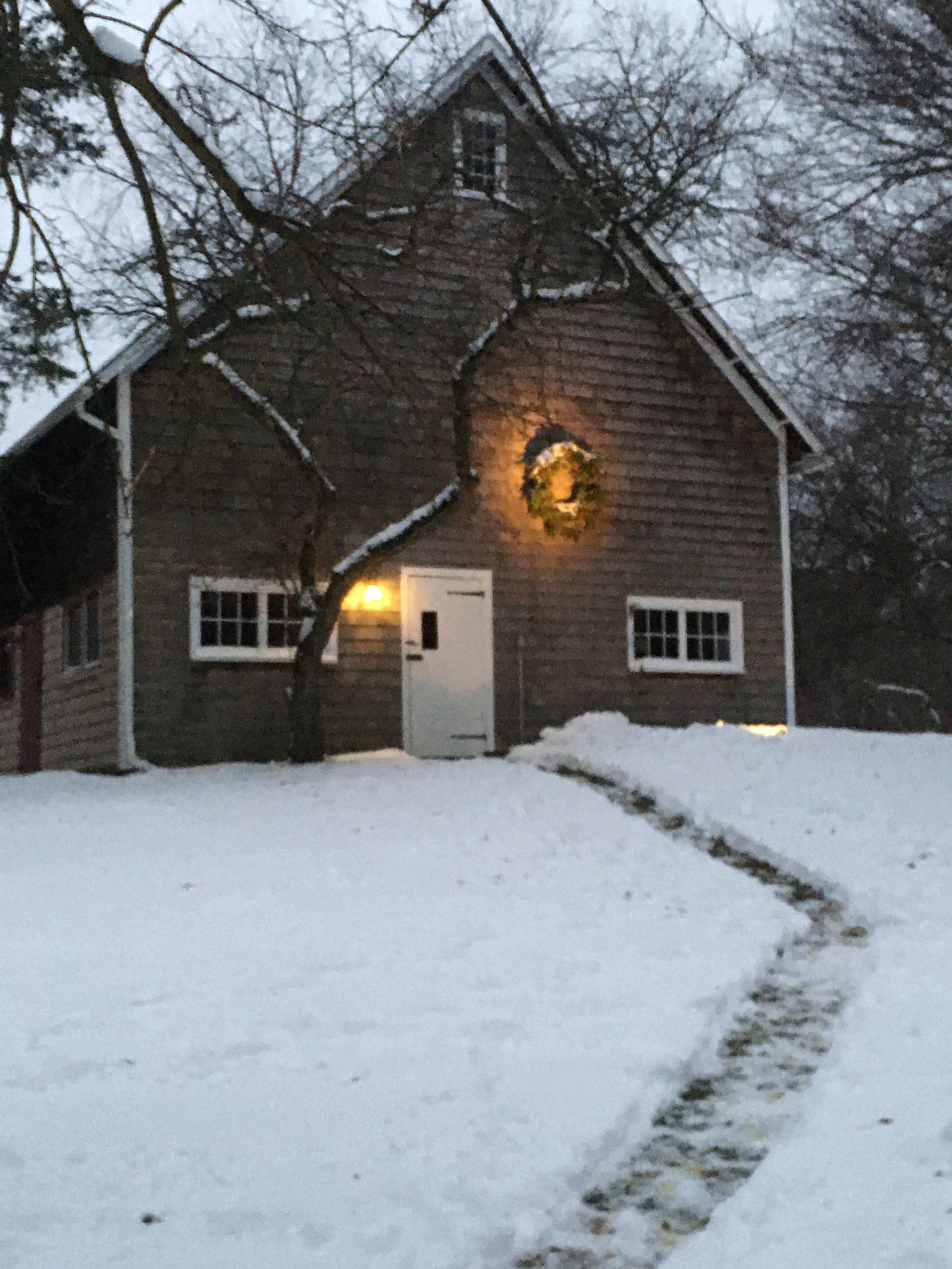 A wooden house with a decorated wreath on the front is surrounded by snow, with a path leading to the door.