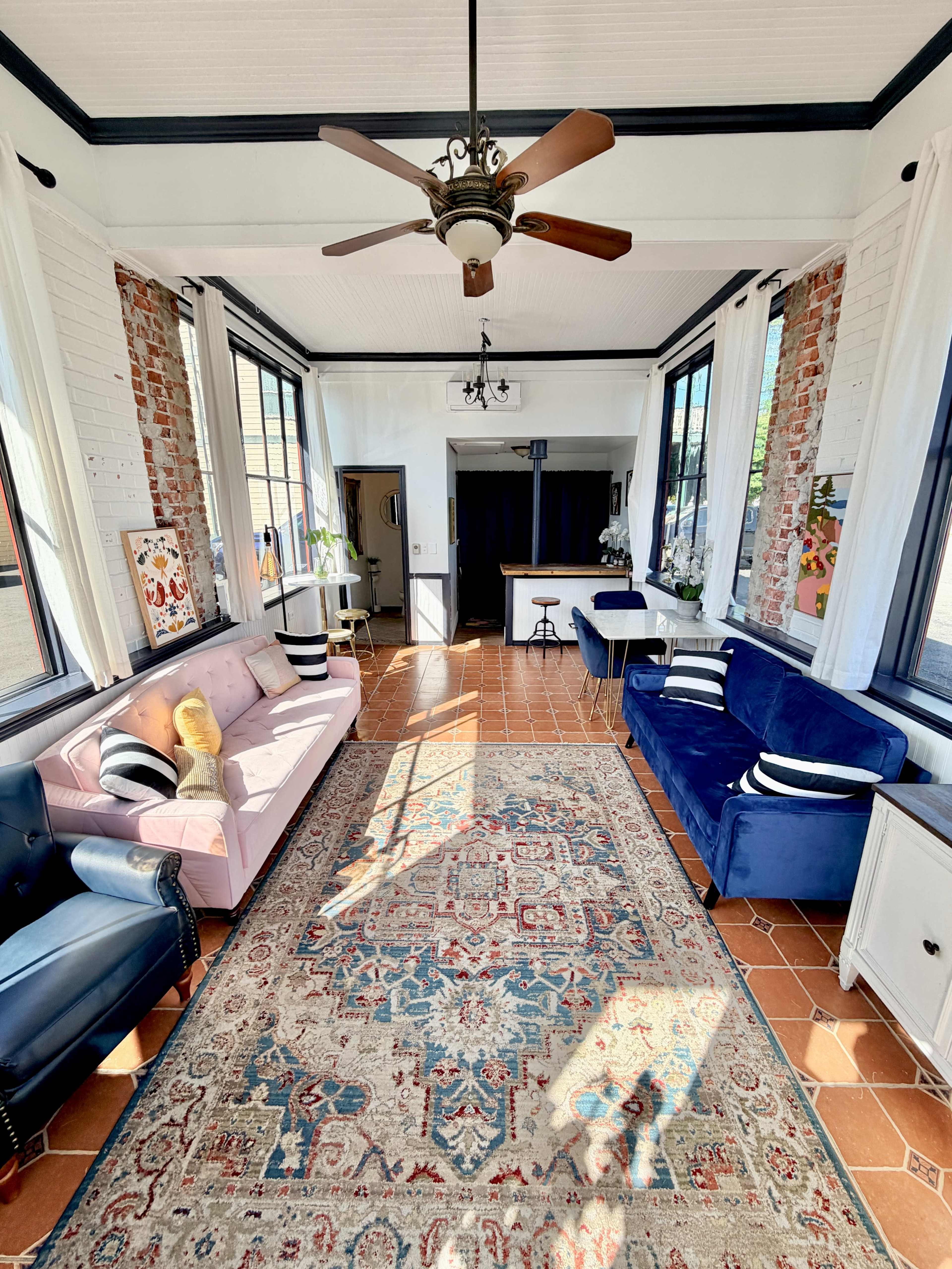 The image shows a bright, sunlit sunroom with a tiled floor, two sofas, a large area rug, and windows lining the walls.