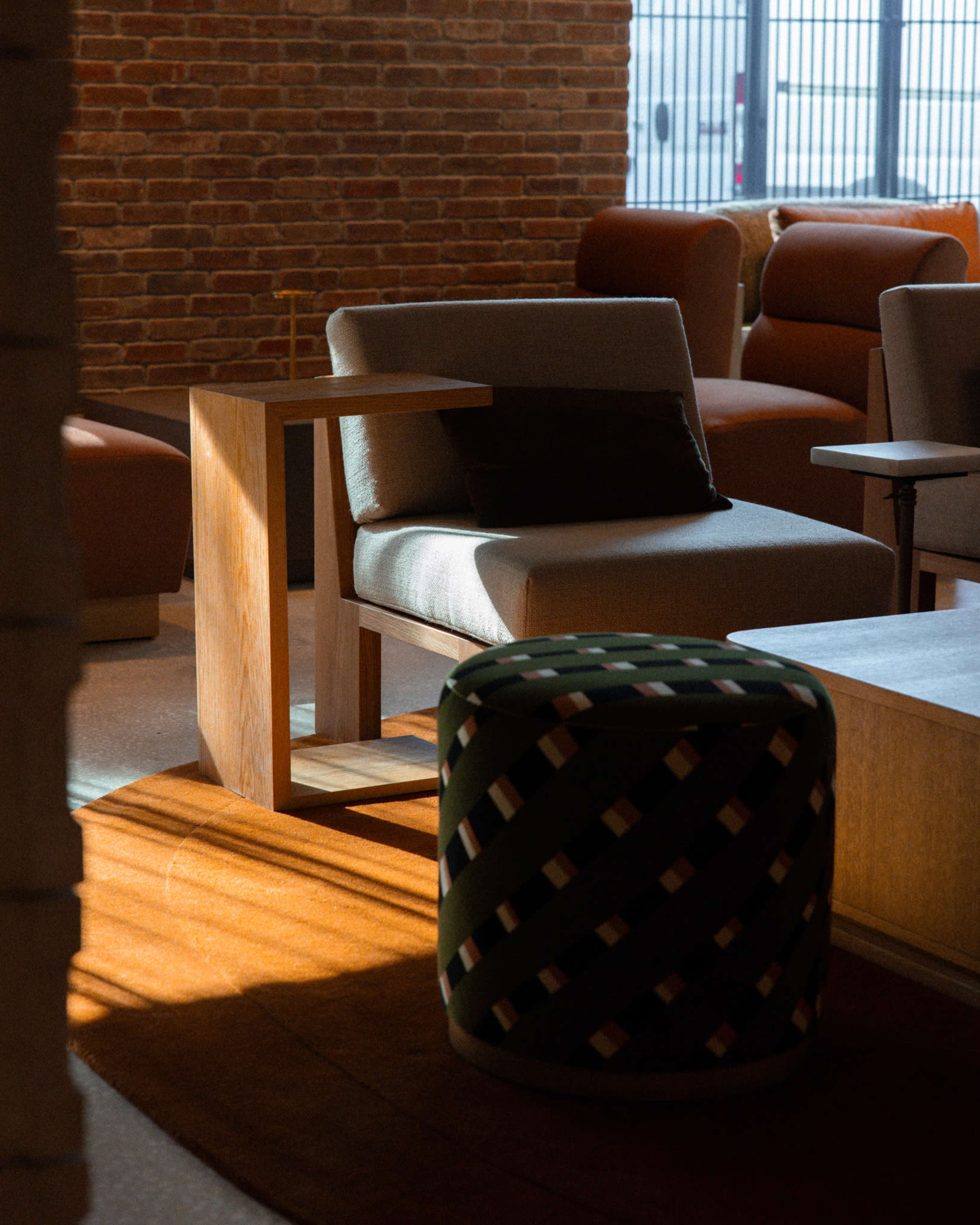 The image features a modern seating arrangement with a wooden armchair and a patterned ottoman in a well-lit interior space.