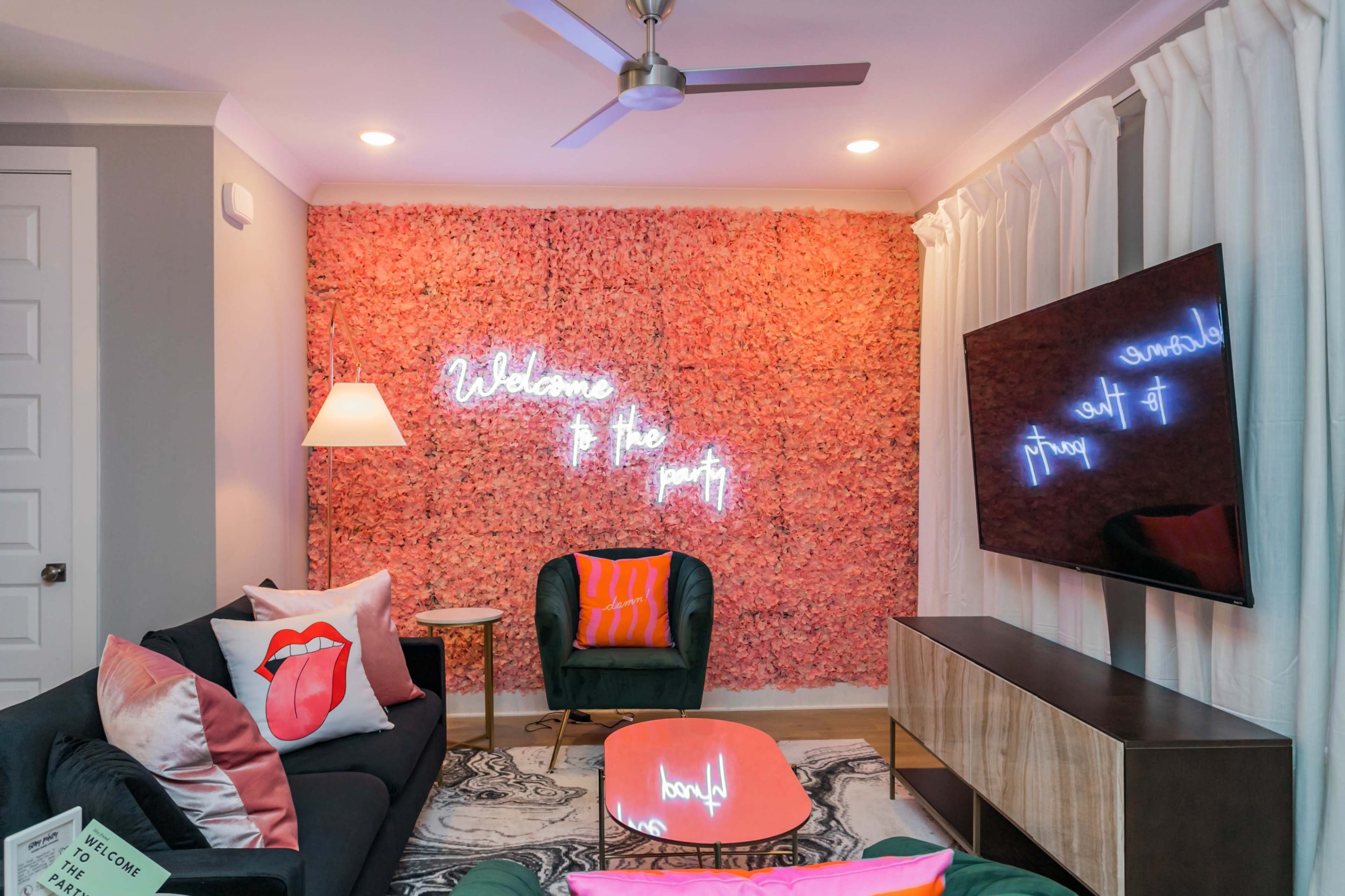 A living room features a pink flower wall with neon signage that reads "Welcome to the party," a green velvet chair, and a modern television.