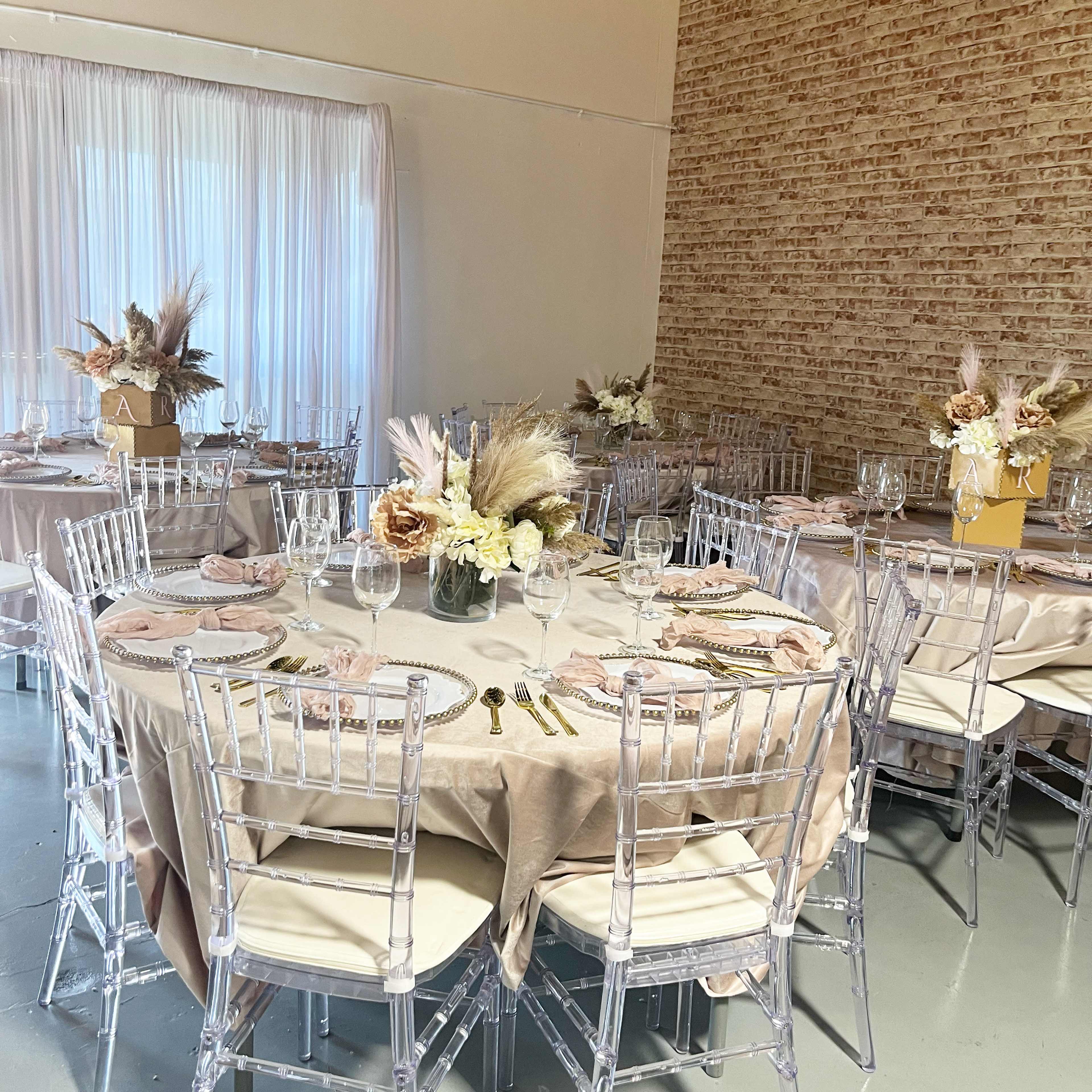 A dining area set up for an event, featuring round tables with beige tablecloths, clear ghost chairs, and floral centerpieces.