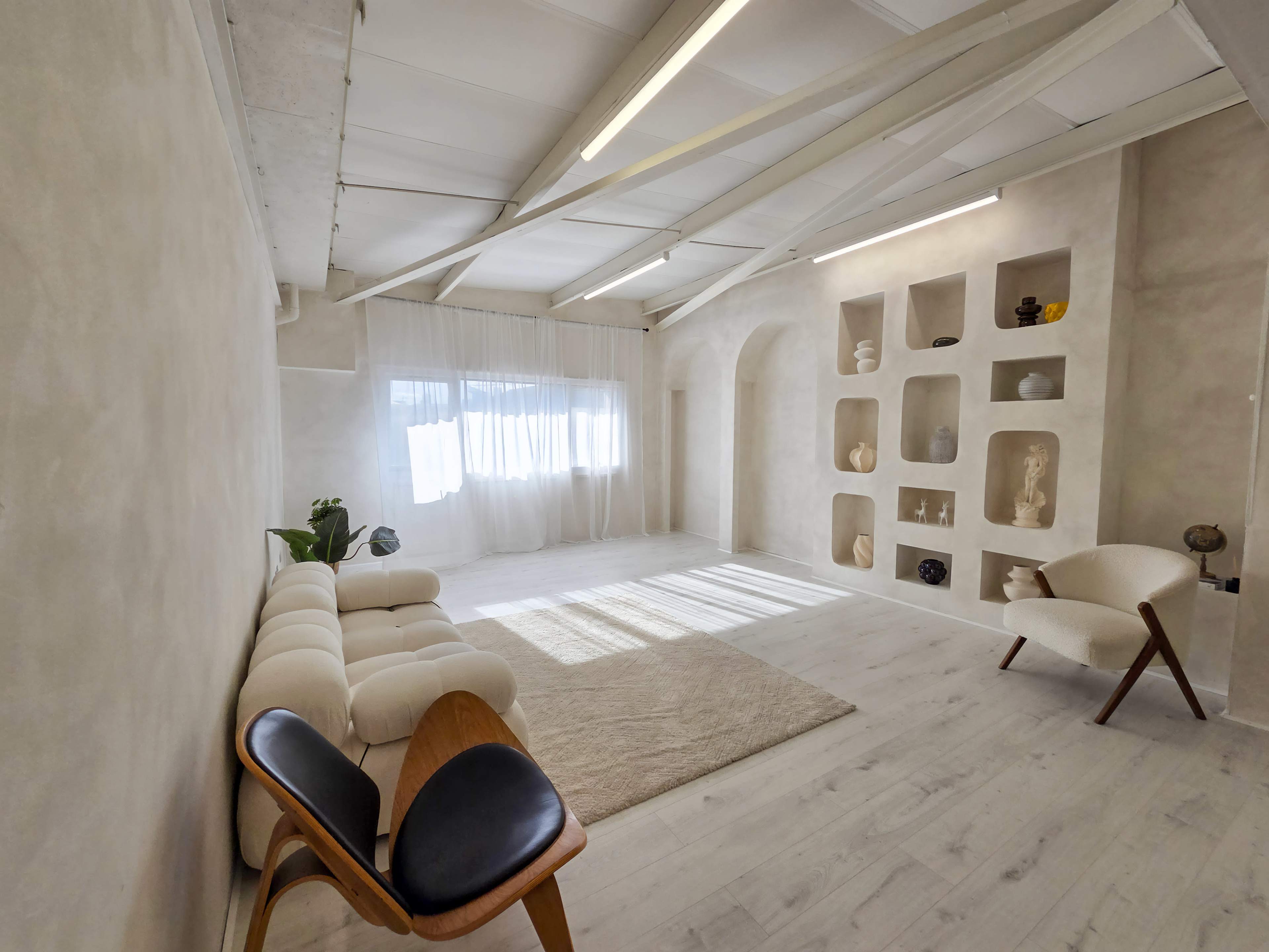 The image shows a minimalist interior space with a white sofa, wooden chair, and a wall featuring built-in shelves displaying various objects.