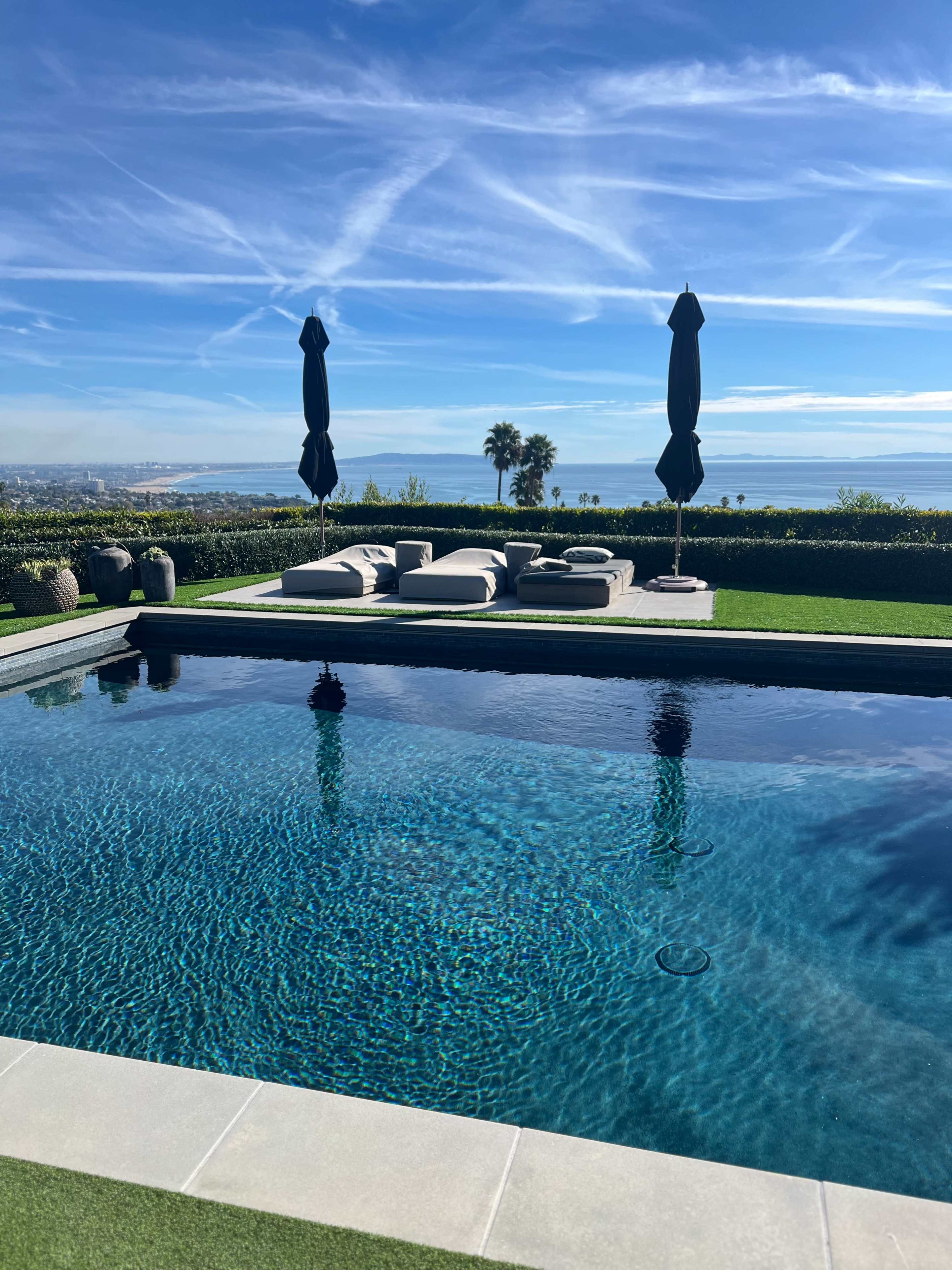 The image shows a modern swimming pool surrounded by lounge chairs and umbrellas, set against a clear blue sky and a distant view of the landscape.
