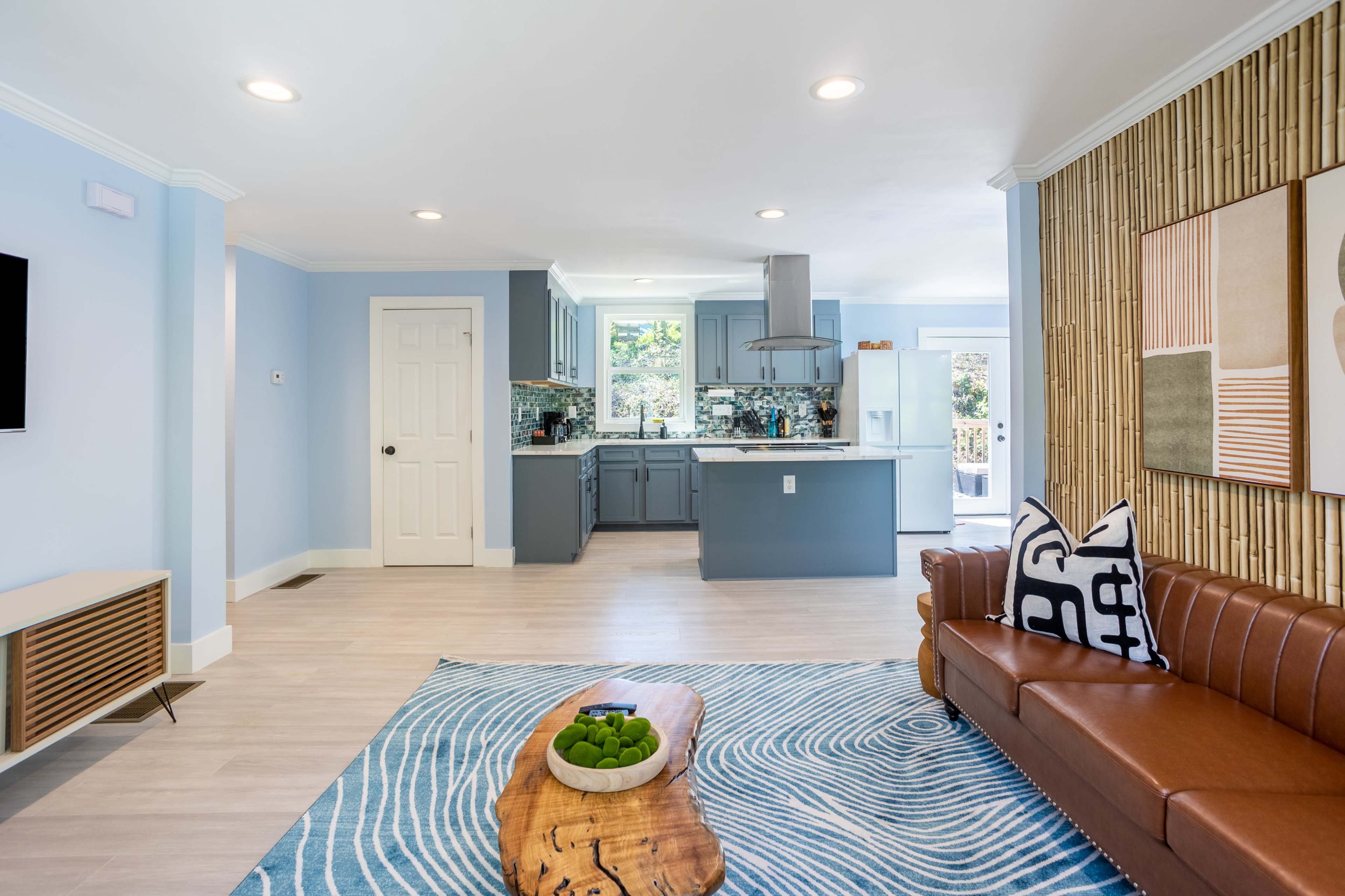 A modern living room with a kitchen in the background, featuring a brown leather sofa, a large area rug, and a wooden coffee table with green fruit.