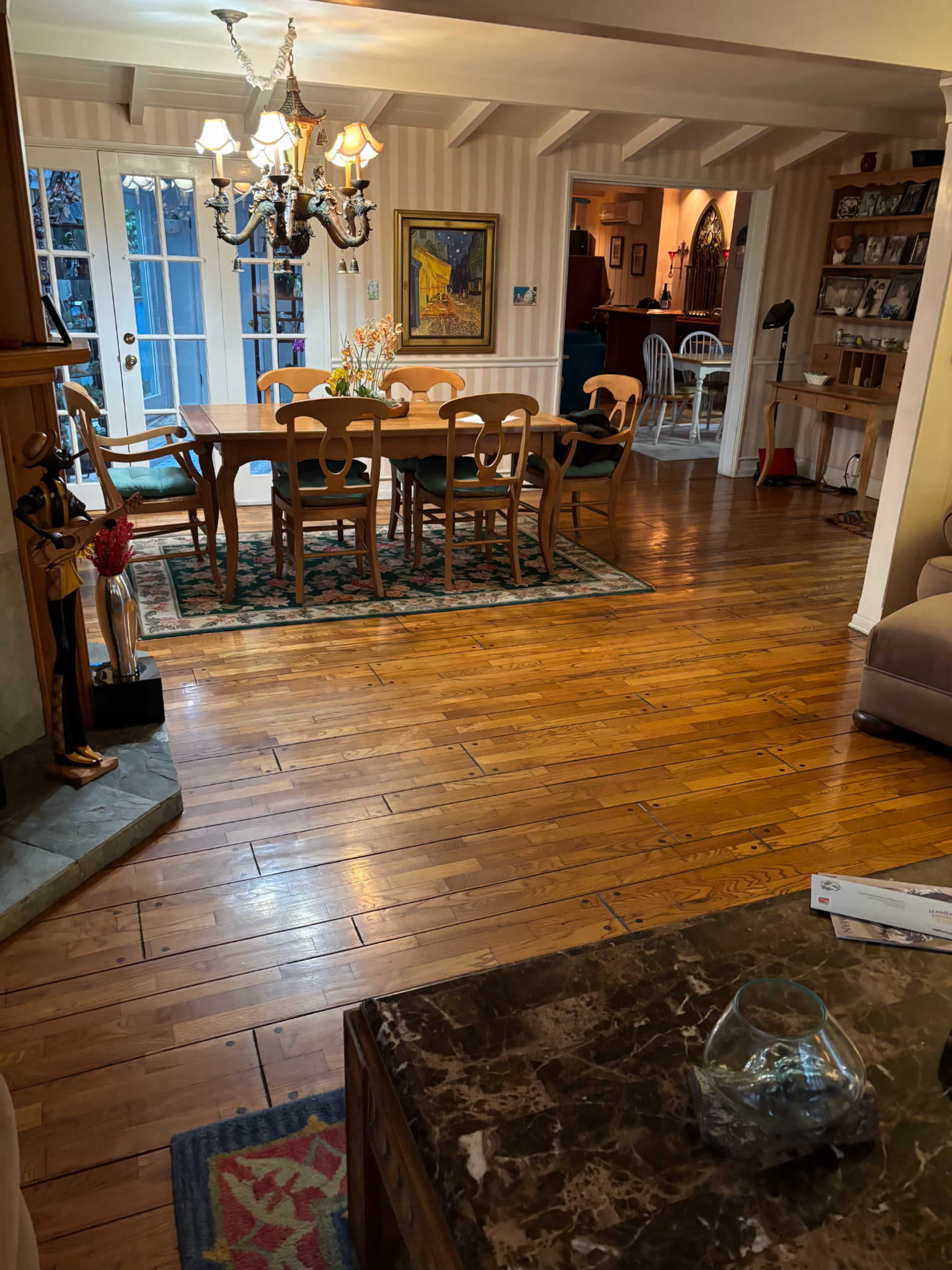 The image shows a dining area with a large wooden table surrounded by chairs, a chandelier overhead, and a view into a living space with warm wood flooring.