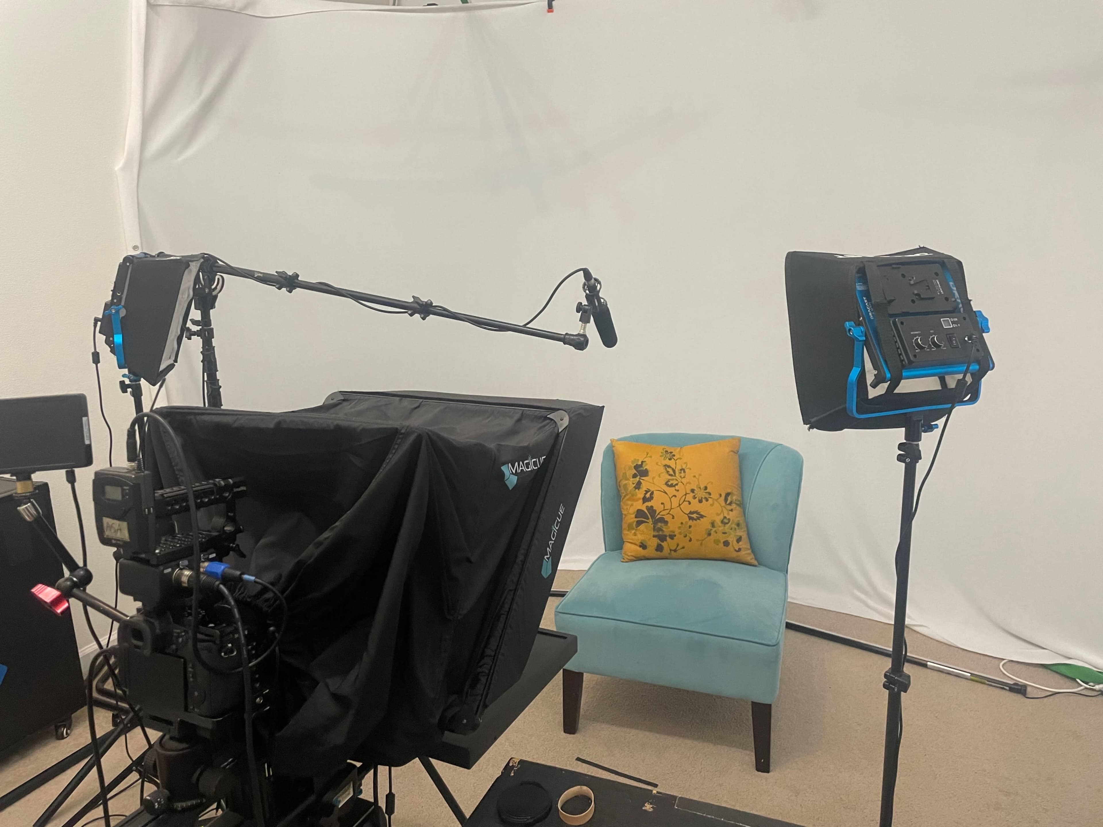A video recording setup features a microphone arm, a camera with a lens hood, and a light on each side, facing a teal chair with a yellow patterned pillow against a white backdrop.