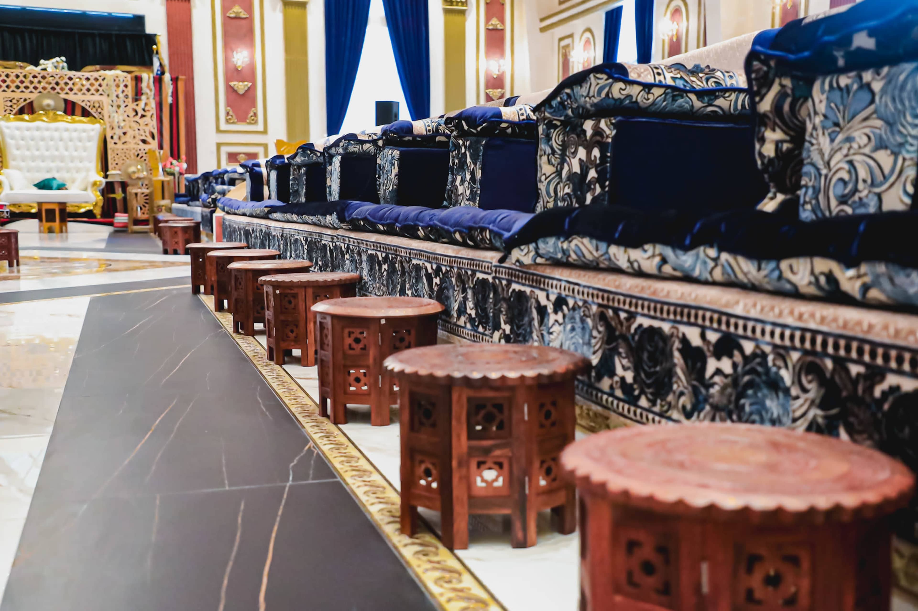 The image shows a lavishly decorated interior with ornate seating arrangements and small wooden stools arranged along a patterned floor.