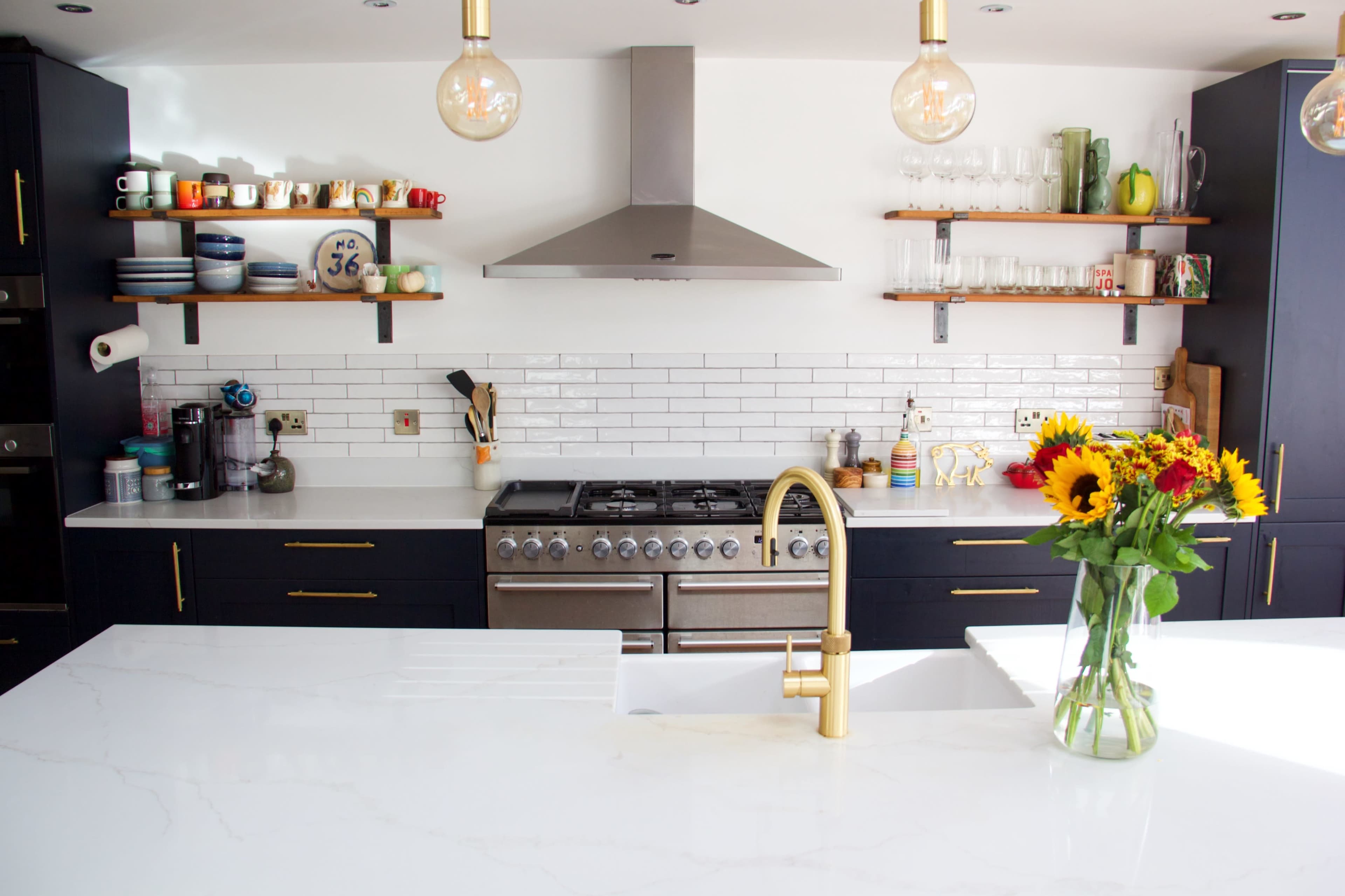 A modern kitchen features a large island with a gold faucet, sleek cabinets, and open shelving displaying dishes and glassware, along with a vase of sunflowers.