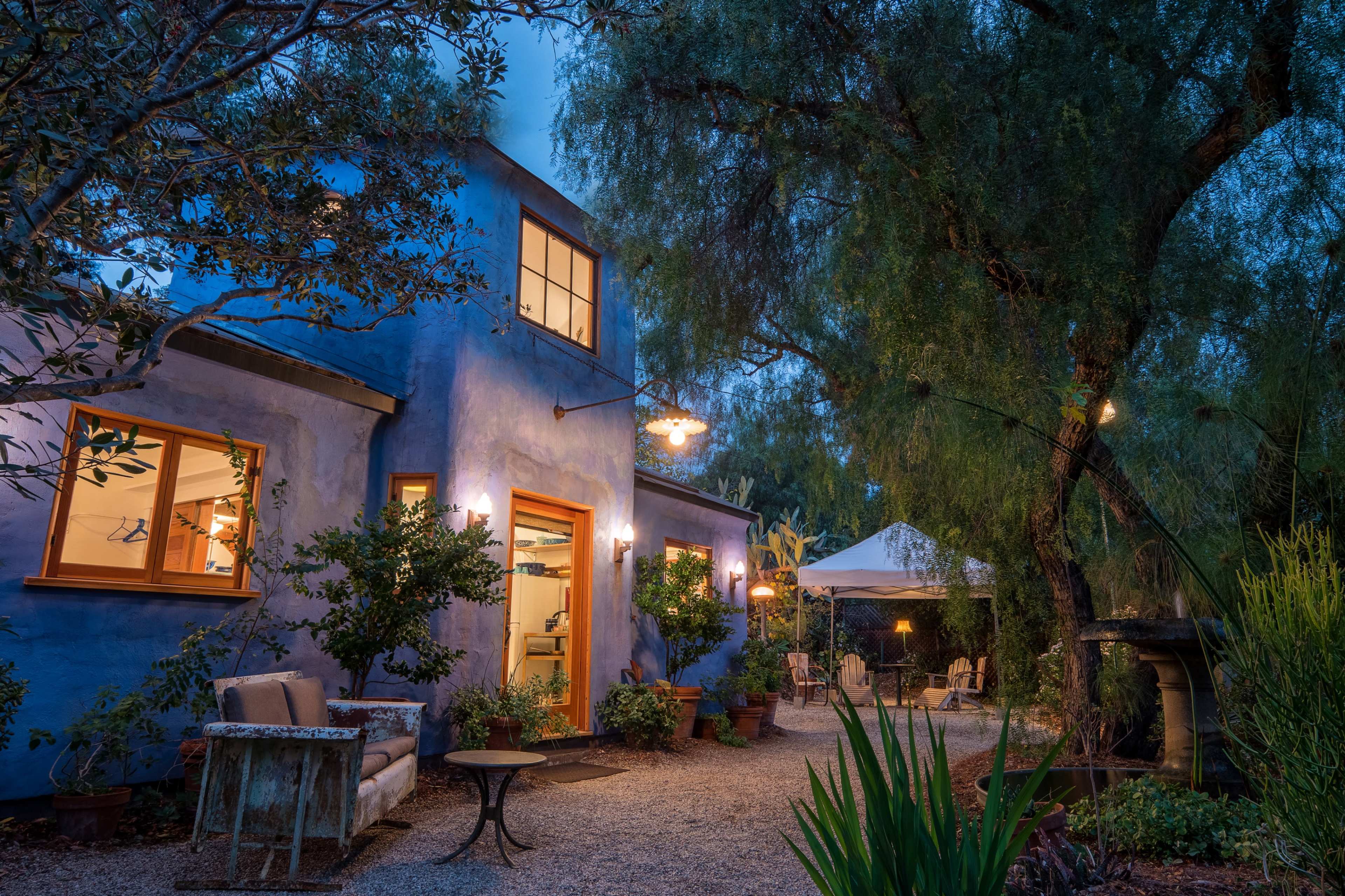 A blue-stucco house with illuminated windows is surrounded by greenery and outdoor seating under a canopy at dusk.