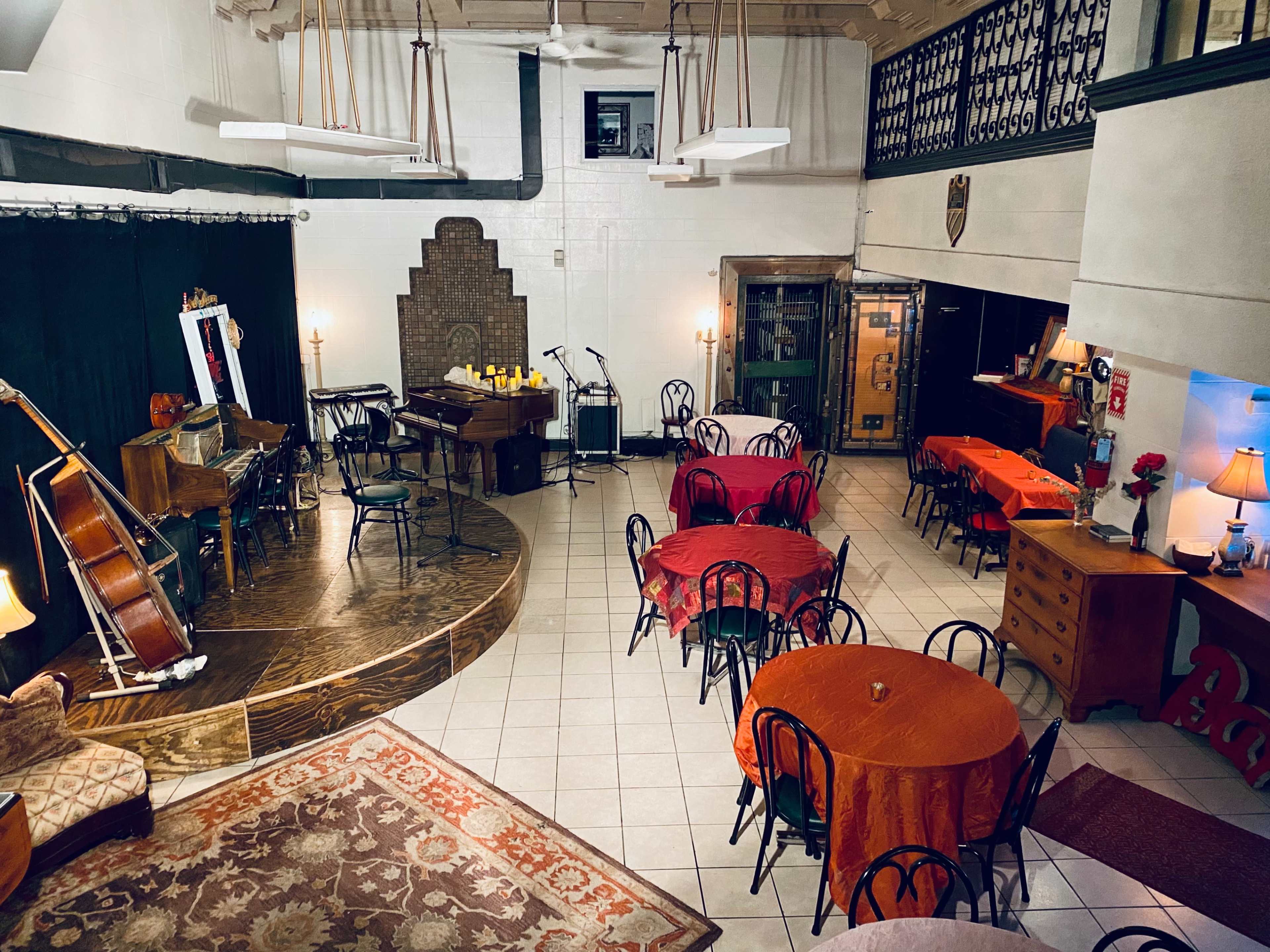 A spacious performance venue with a wooden stage, musical instruments, and several round tables set with orange tablecloths.