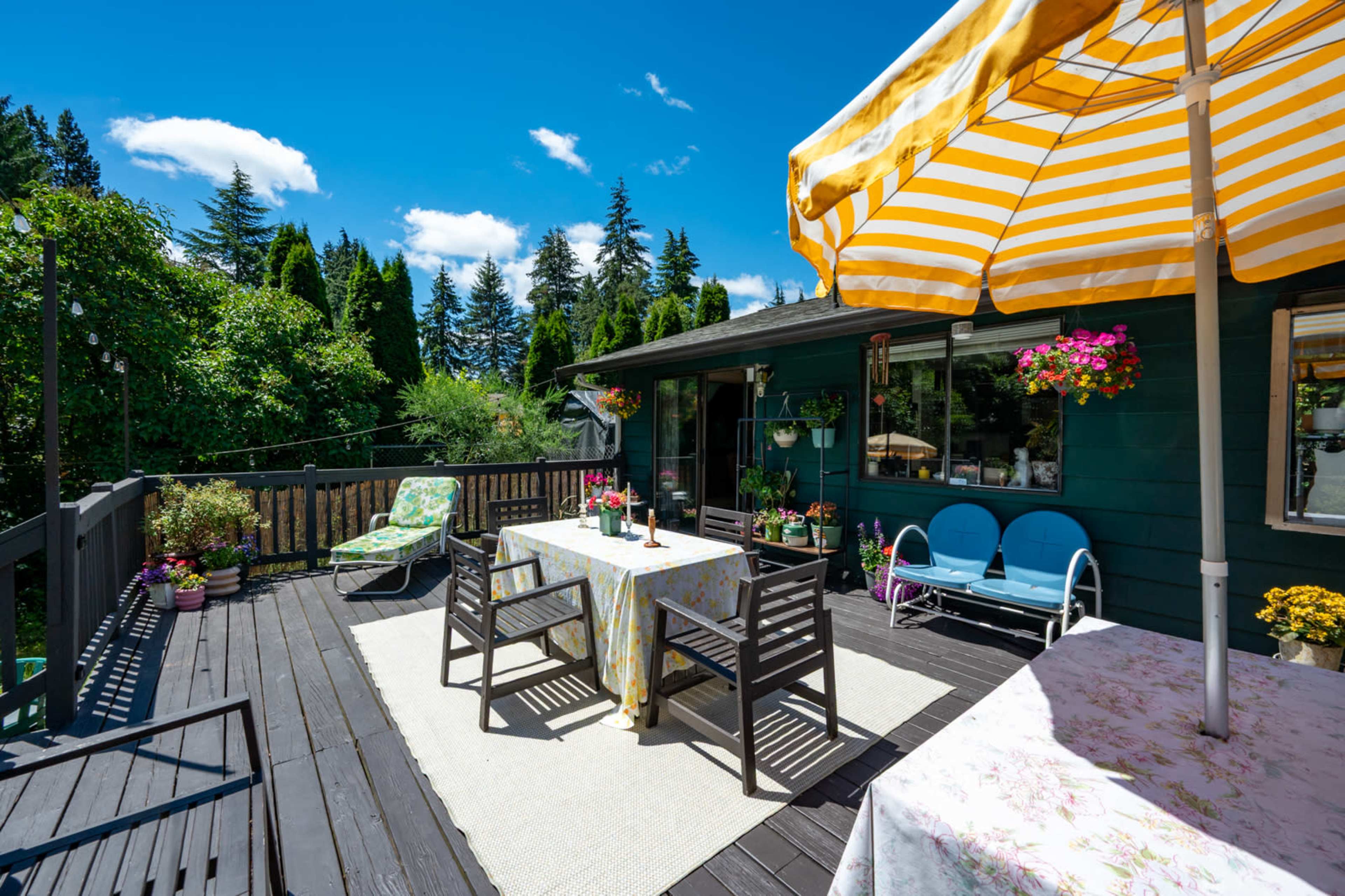The image shows a bright outdoor deck area with two tables, chairs, and a striped umbrella, surrounded by greenery and potted flowers.