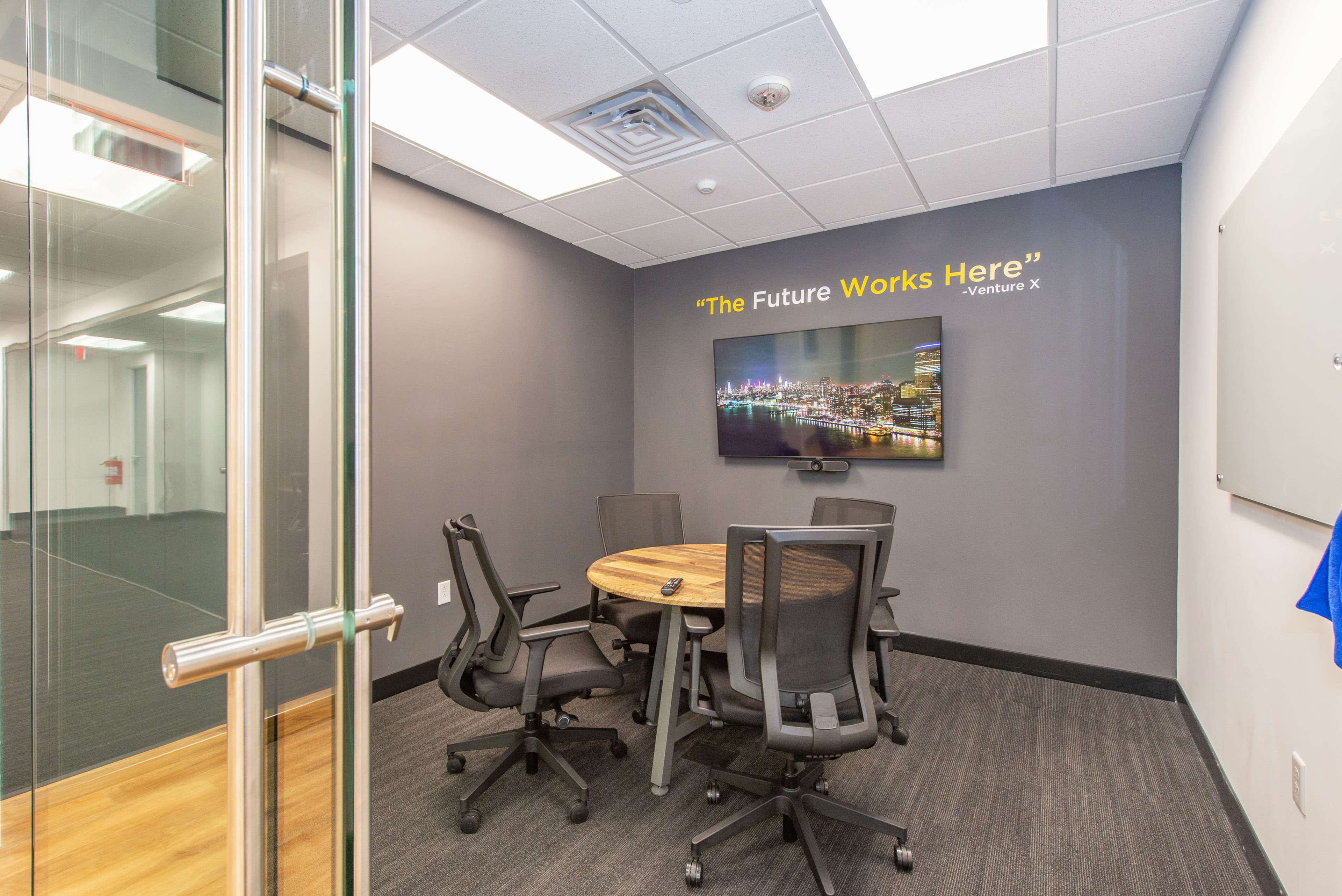 A modern conference room features a round wooden table surrounded by black chairs, with a television displaying the text "The Future Works Here" on the wall.