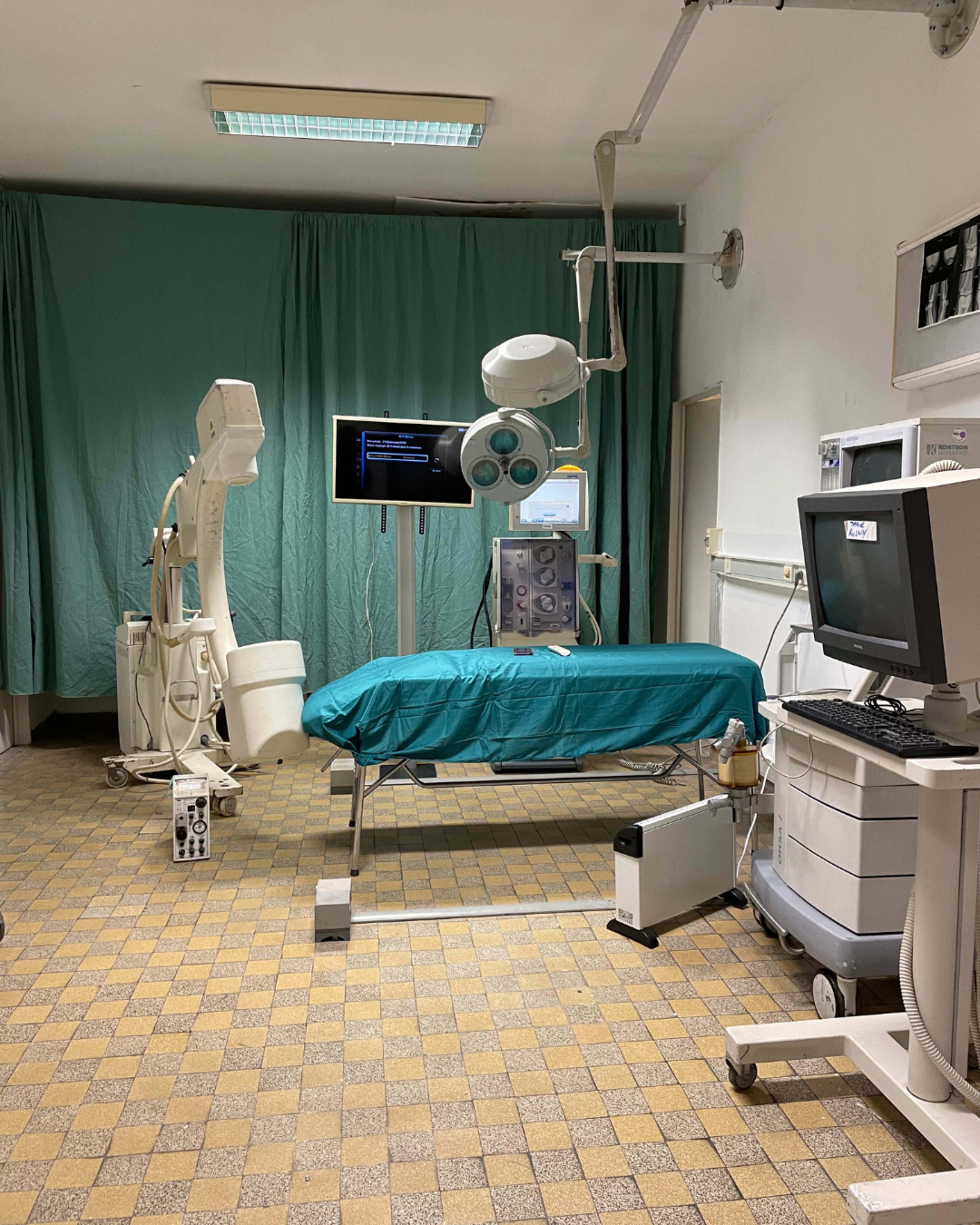 A medical examination room featuring a bed with a teal cover, various medical equipment, and a computer workstation.