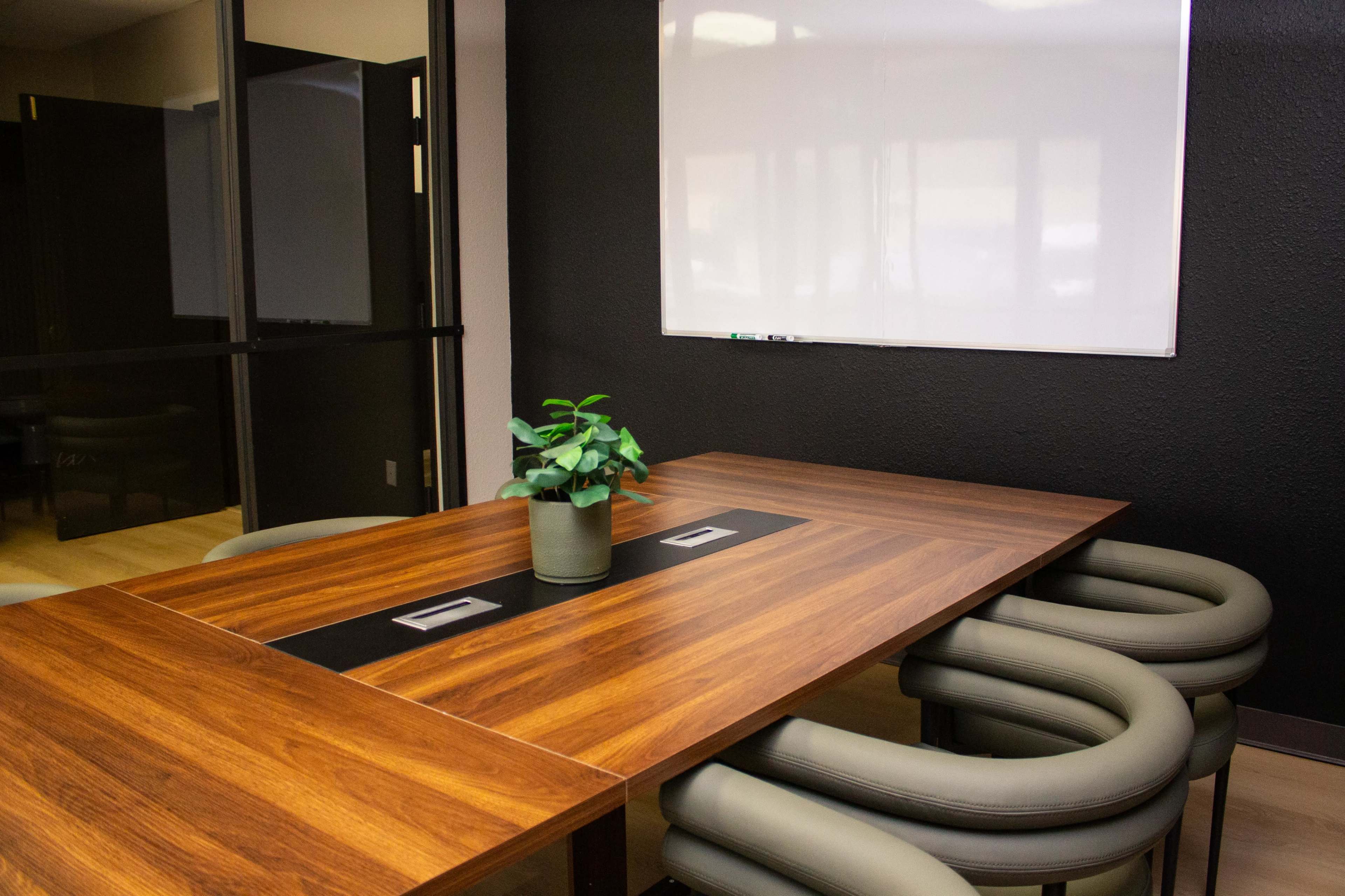 The image shows a modern conference room featuring a wooden table with a central planter and padded chairs, alongside a whiteboard mounted on a dark wall.
