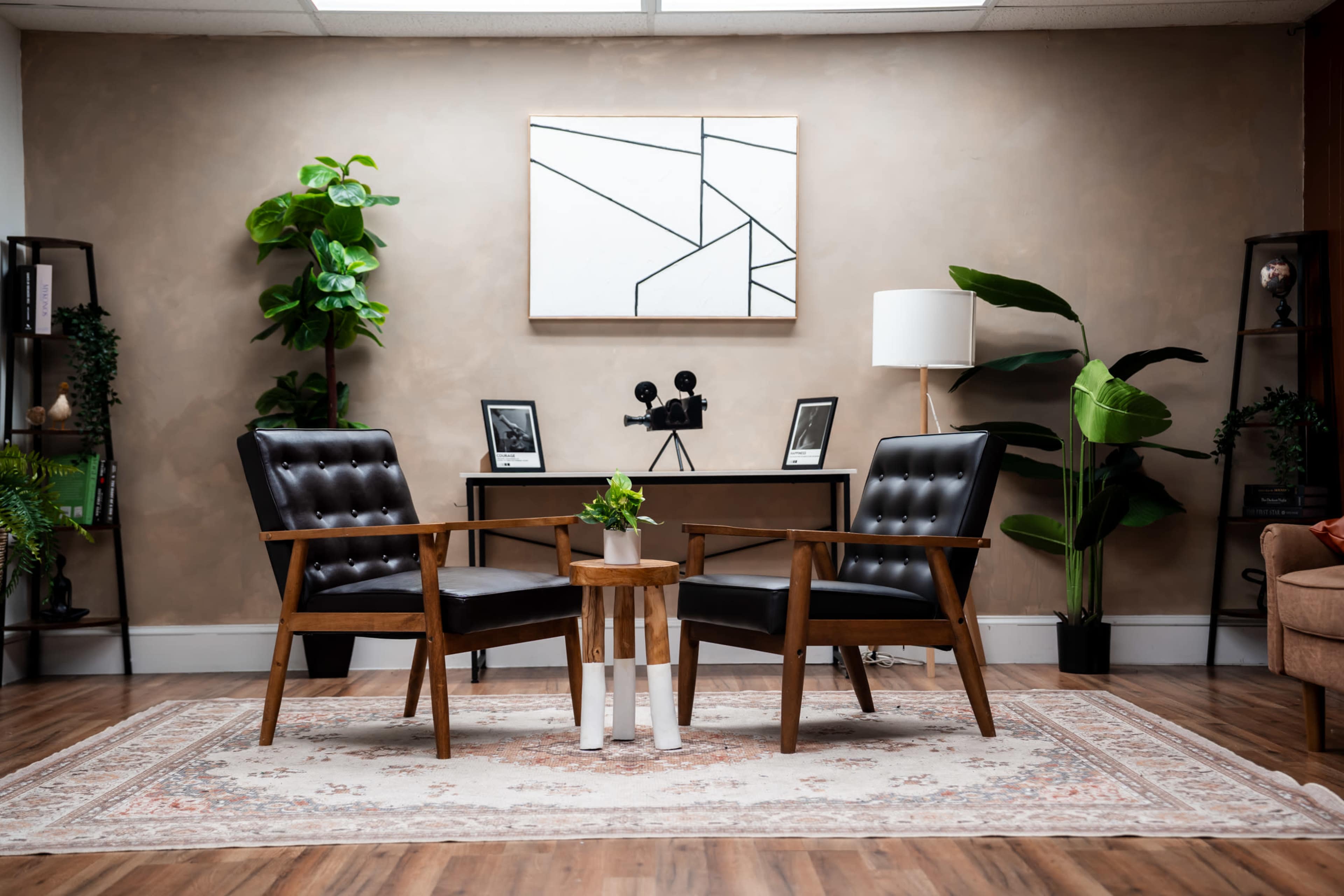 The image depicts a modern interior scene featuring two wooden armchairs with black cushions facing a small round table, surrounded by decorative plants and framed pictures on the wall.