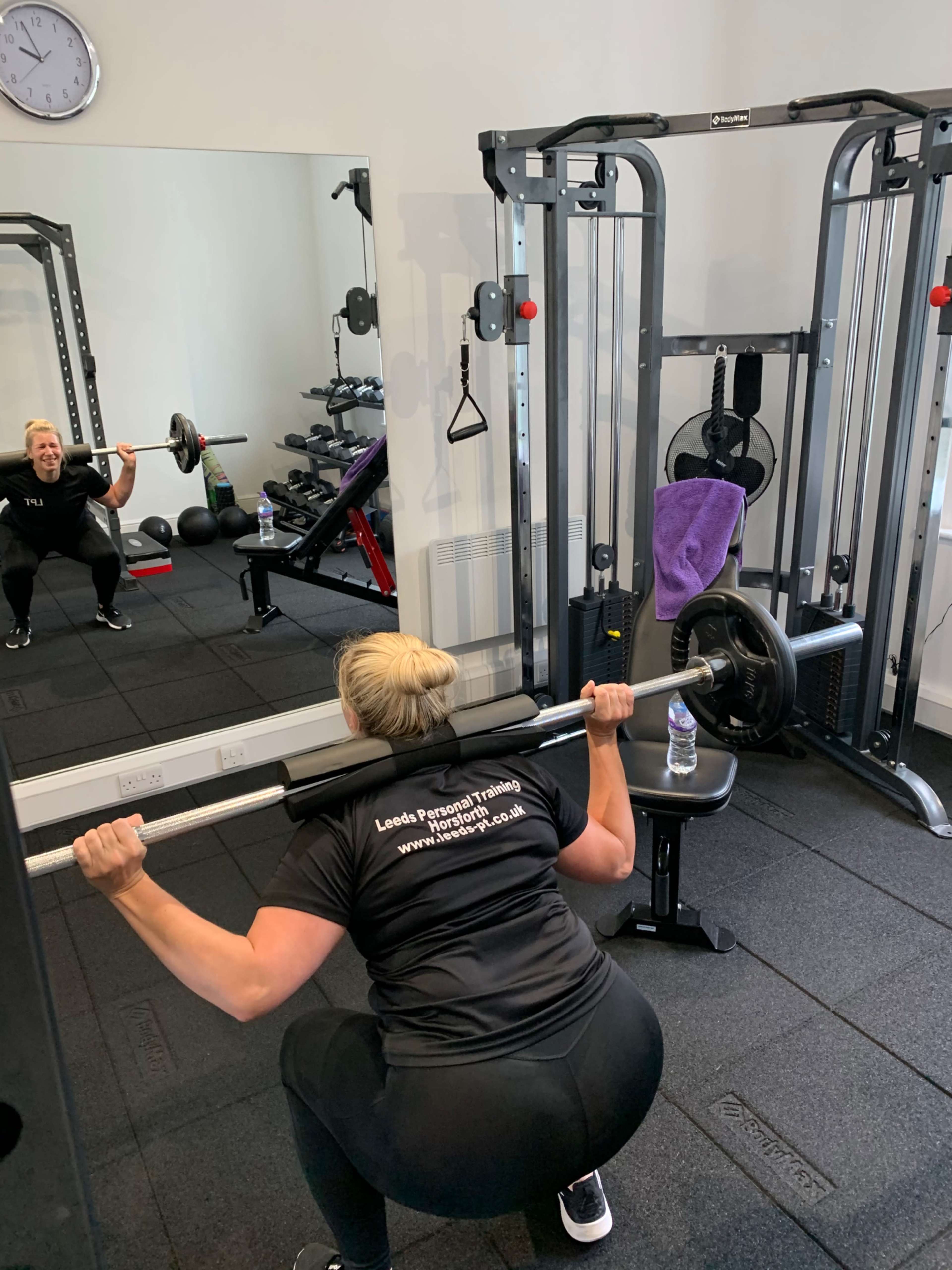 A person is performing a squat with a barbell in a gym, with a mirror reflecting the scene and exercise equipment in the background.