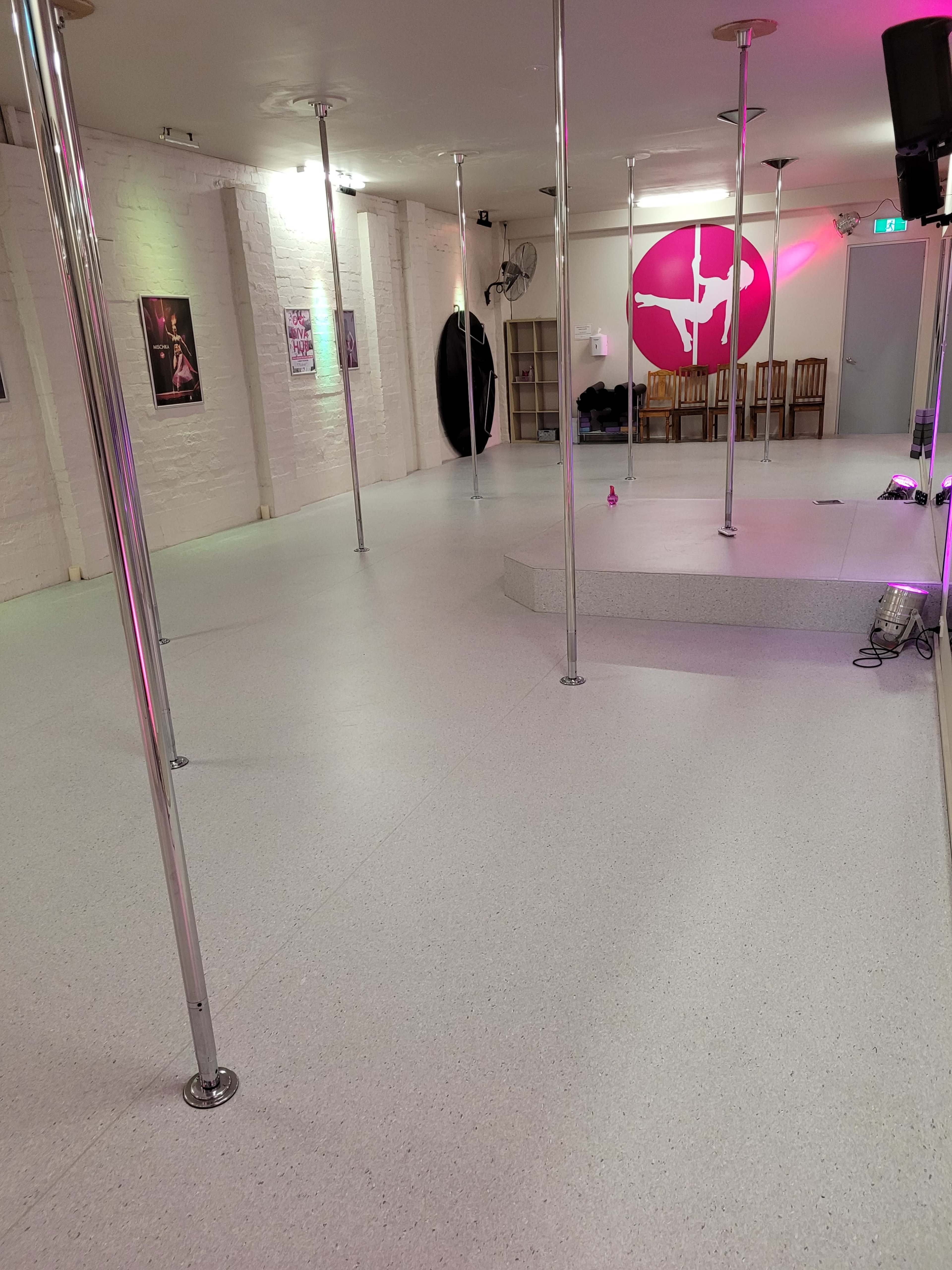 The image shows a dance studio with multiple shiny poles, a circular stage, and colorful lighting.